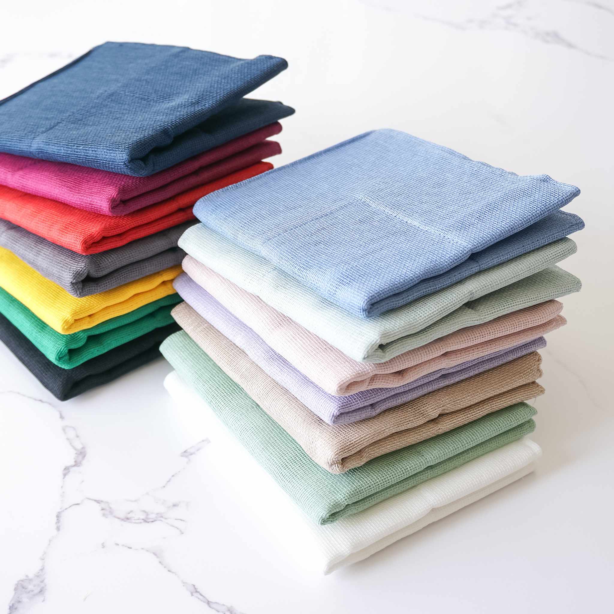 Stacks of folded Japanese dishcloths in various solid colors including blue, red, yellow, green, beige, and white, displayed on a marble surface.