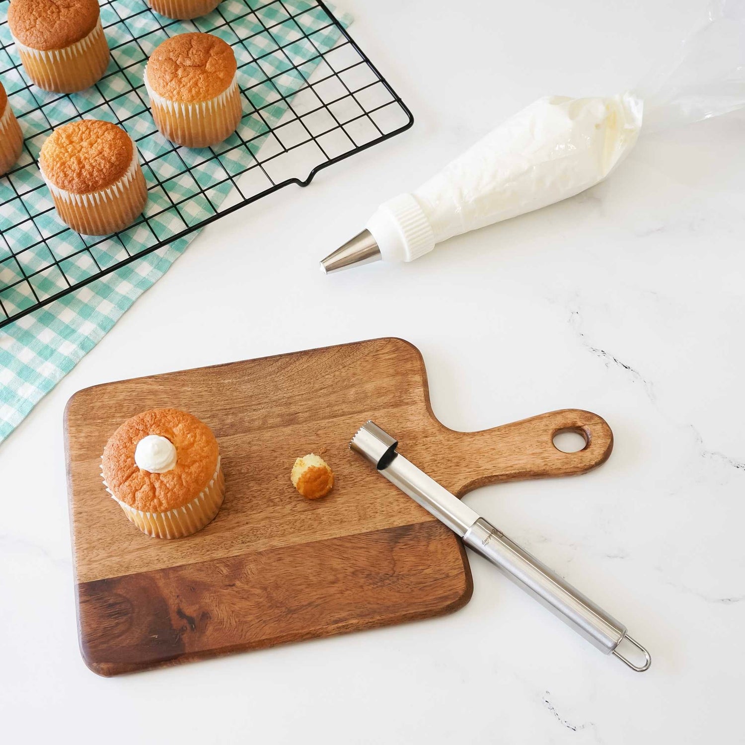 A stainless steel cupcake corer sitting on a wooden cutting board with a cupcake and its filling being demonstrated using the corer. In the background, there are cupcakes on a cooling rack and a piping bag with frosting.