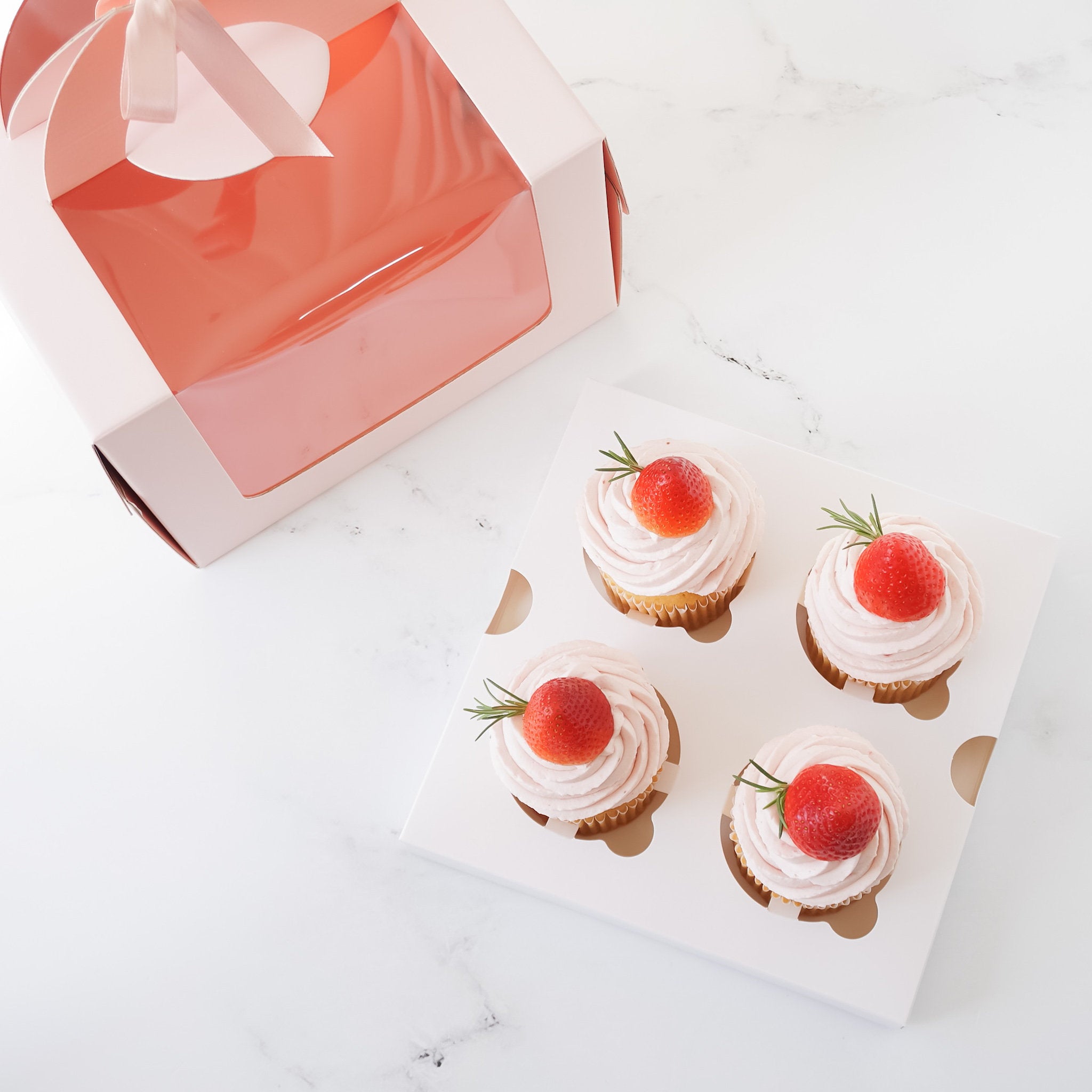 Four cupcakes with white frosting and a strawberry on top, presented in a glossy white cupcake insert.