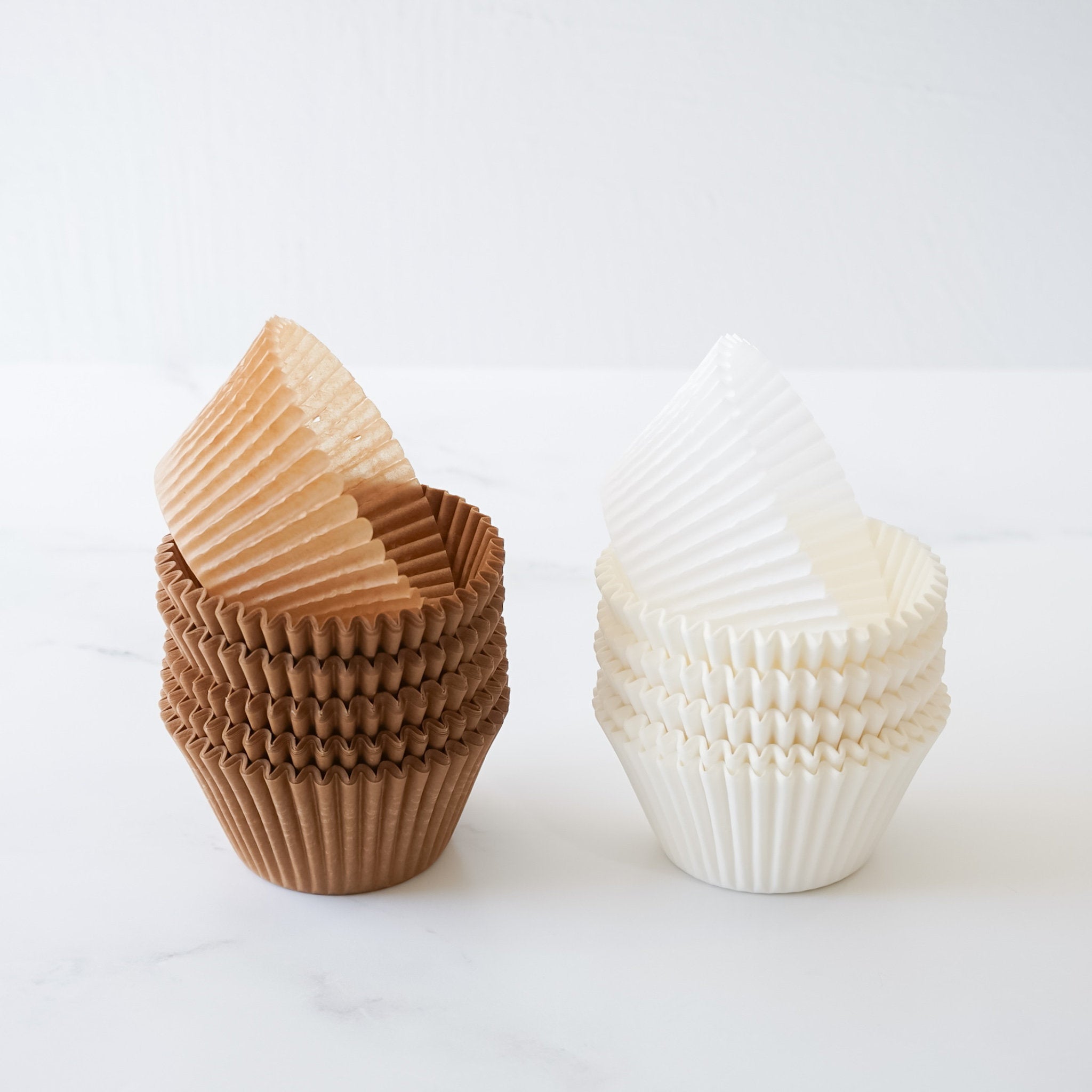 Two stacks of brown and white cupcake liners. The brown liners are stacked on top of each other and the white liners are stacked on top of each other. The liners are both made of paper and have a fluted design.
