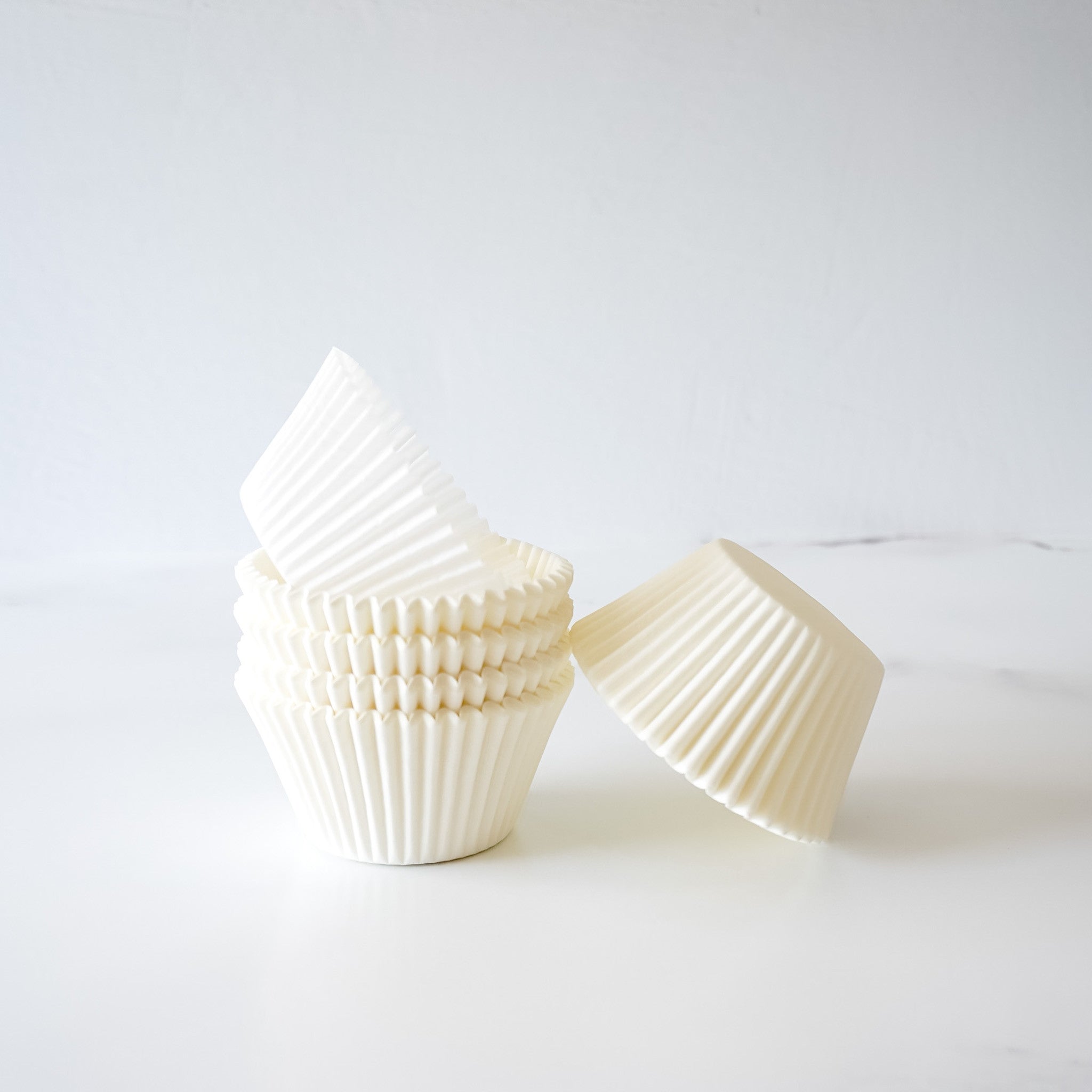 A stack of white cupcake liners, with one liner laying on its side. The liners are made of paper and have a fluted design. The liners are a popular baking supply for making cupcakes and muffins.
