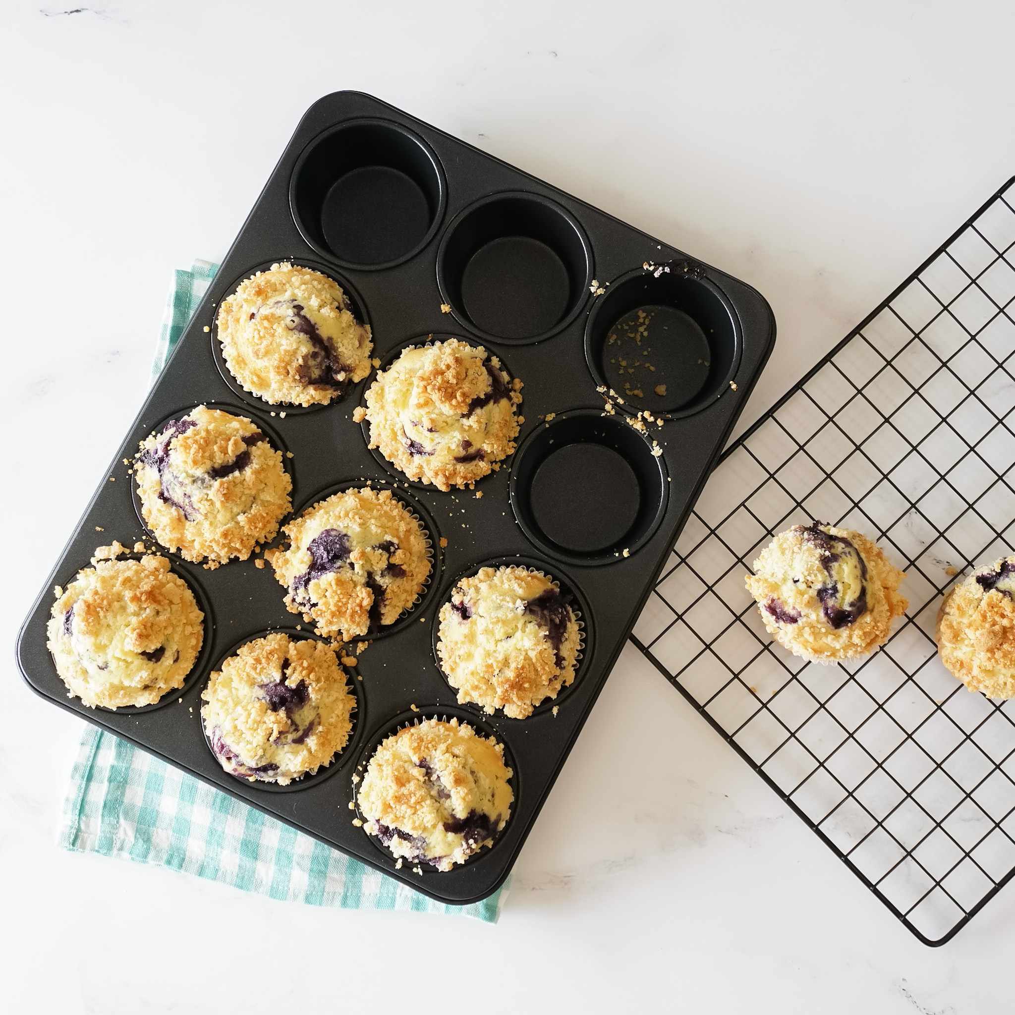 A black cupcake pan with 8 cupcakes inside. The cupcakes are topped with a crumb topping and have blueberries inside. The muffins are on a white surface with a green and white checkered cloth underneath. There are two additional muffins on a black wire rack.