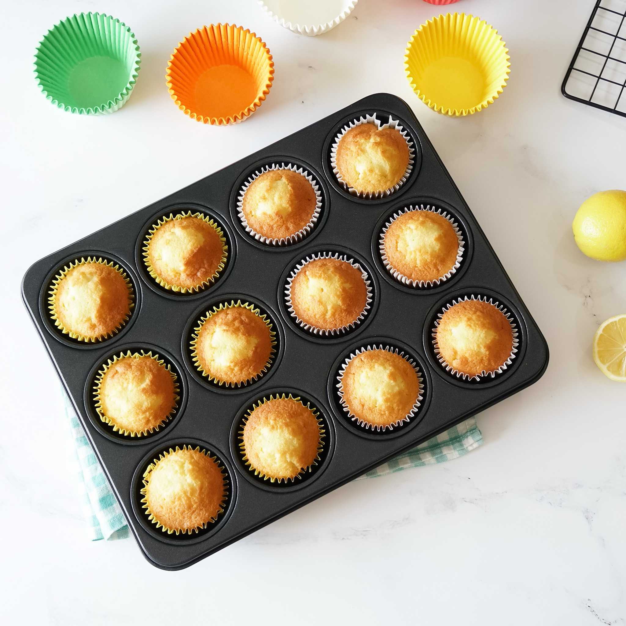 A black muffin tin filled with twelve golden brown cupcakes. The tin is on a white surface with a green and white checkered napkin underneath. There are three empty cupcake liners in the background, one green, one orange, and one yellow.