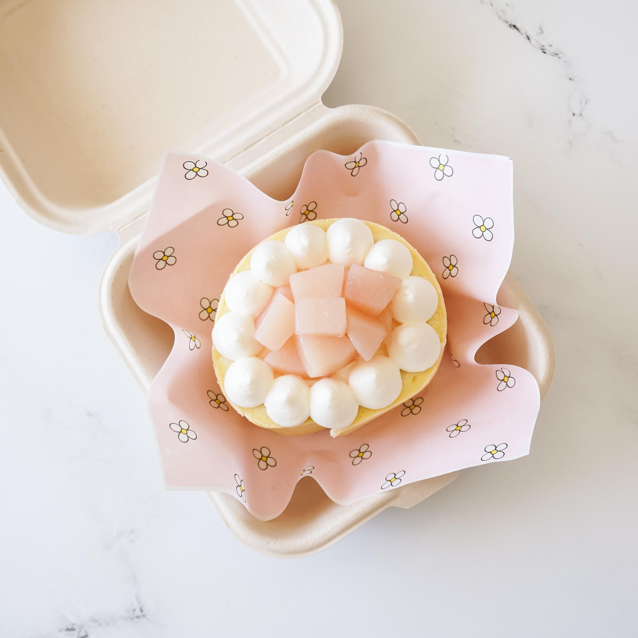 A small deli paper with a pink daisy design, partially liner a carry-out box with a cake on top.