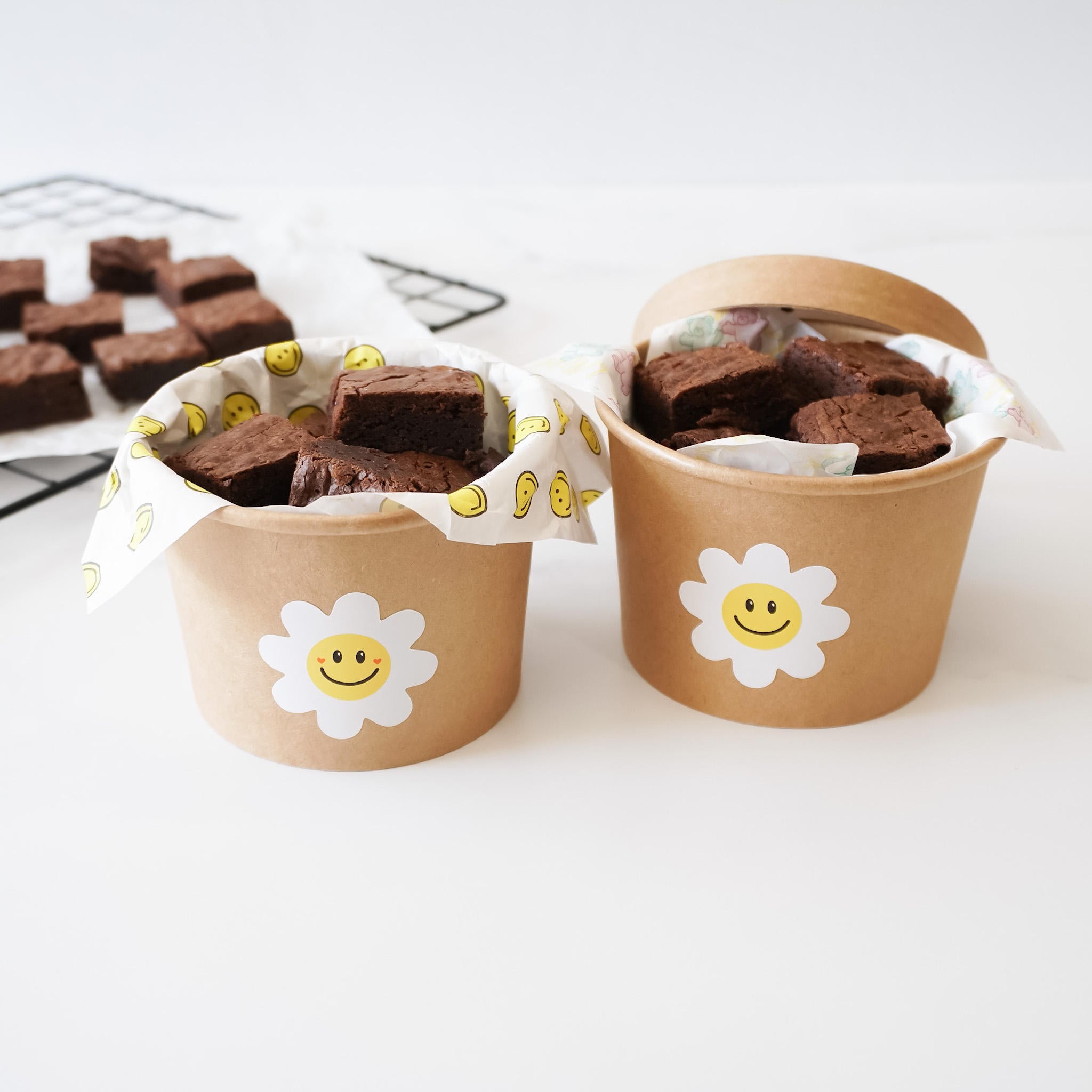The image shows two round brown paper containers with white smiley daisy stickers, filled with chocolate brownies.