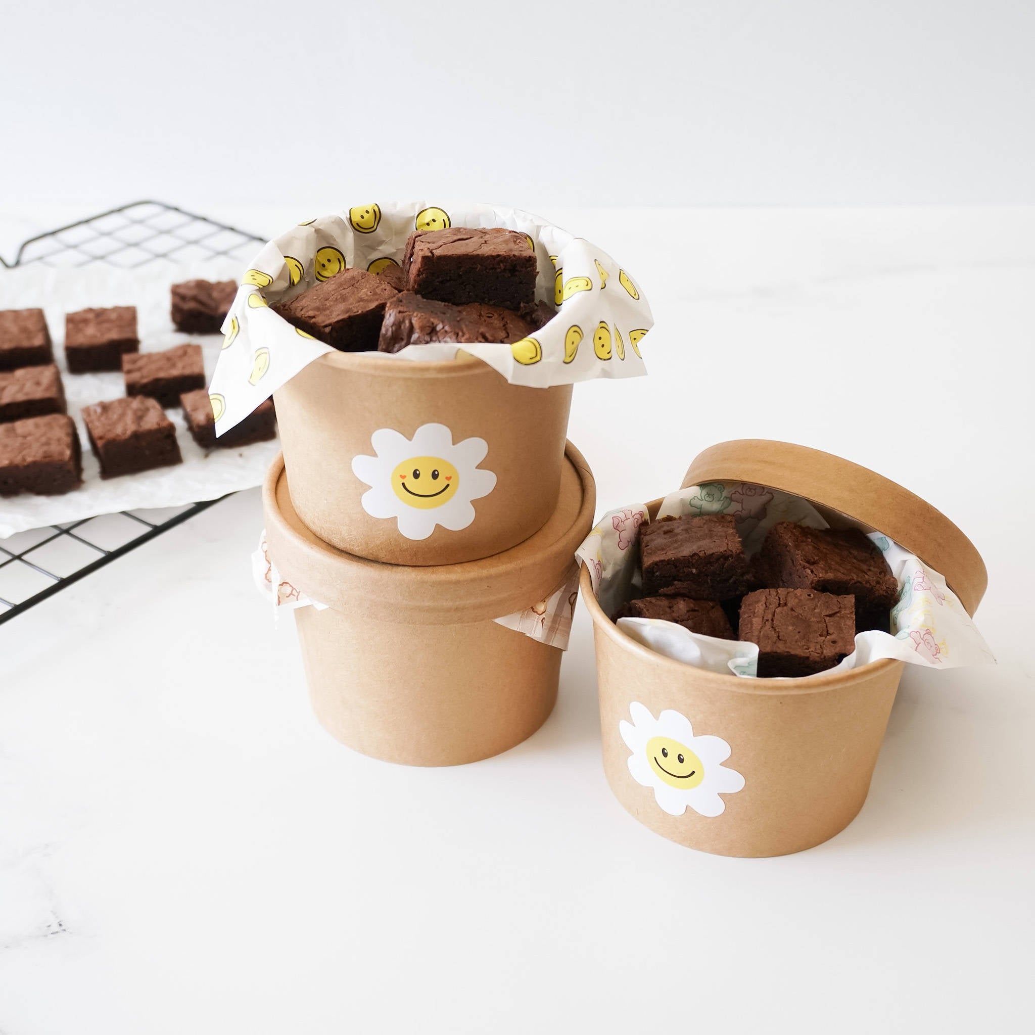 Three brown paper cups with lids, each containing chocolate brownies. The cups are decorated with white daisy stickers with yellow smiley faces. The cups are stacked on top of each other.