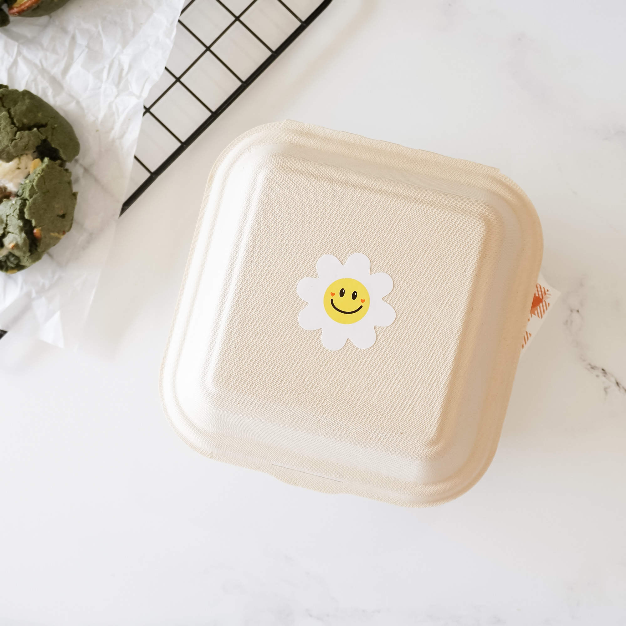 A white, square, cardboard container with a white daisy sticker with a yellow smiley face in the center.