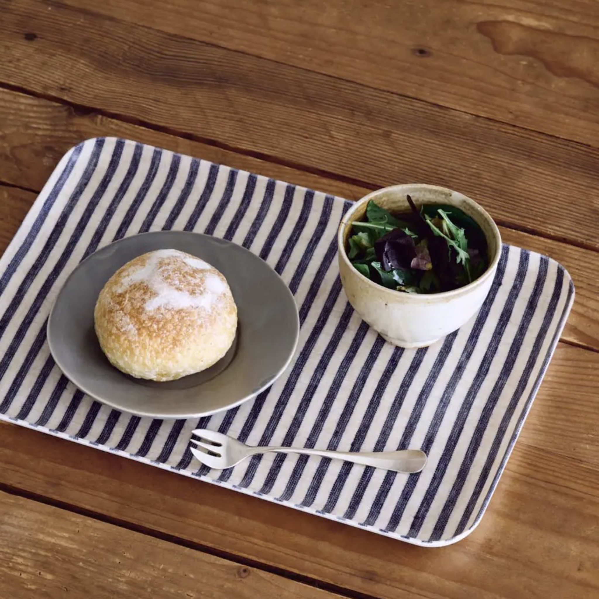 Bun on a plate with a bowl of greens on a striped tray over a wooden table