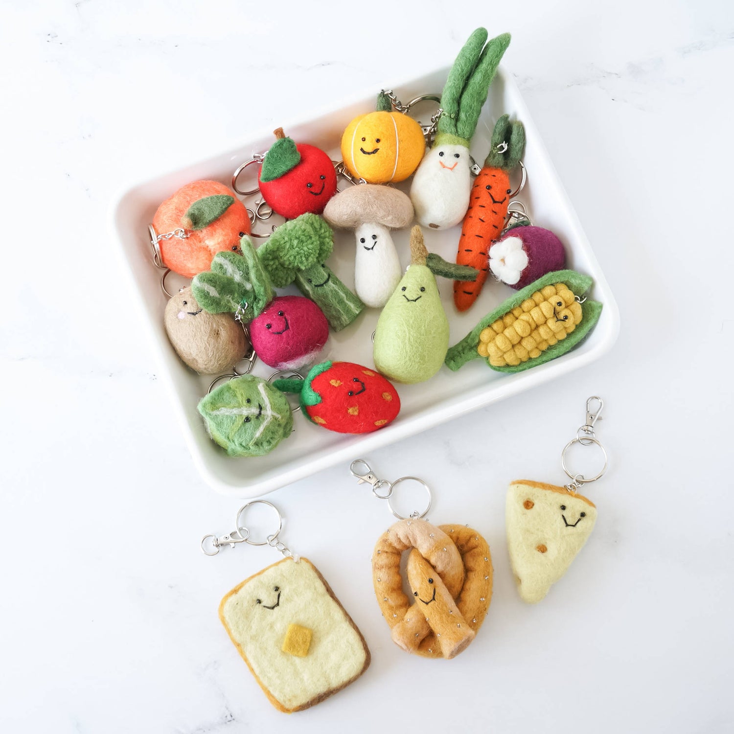 A collection of handcrafted felt keychains in the shapes of various fruits, vegetables, and bread items, displayed on a white plate and marble surface.