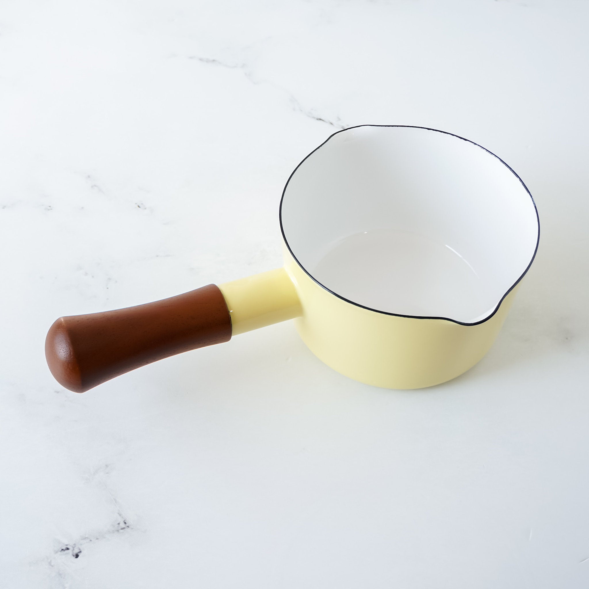 A butter yellow enamel-coated steel milk pan with a wooden handle, displayed against a marble background.