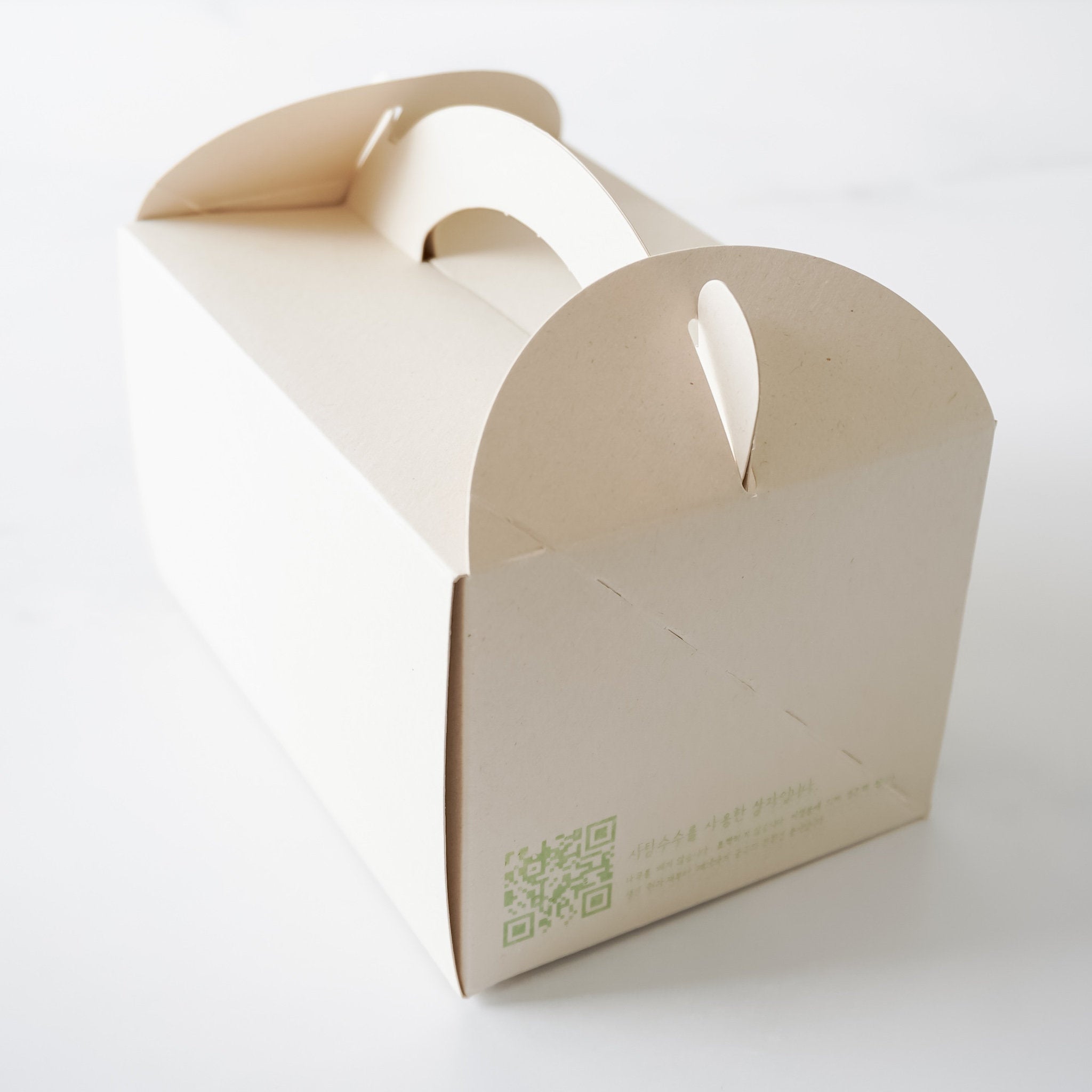 A light beige cardboard box with a handle on the top. The box has a green QR code and text in Korean on the side.