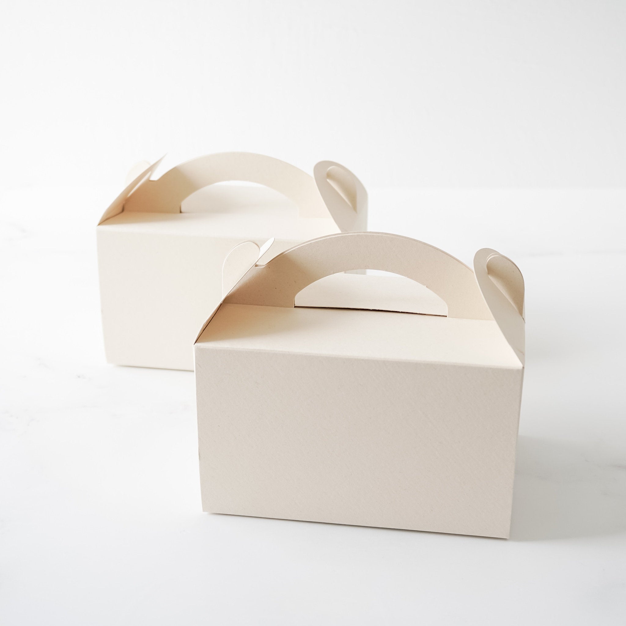 Two light beige cardboard boxes with cut-out handles. The boxes are empty and sitting on a white surface.