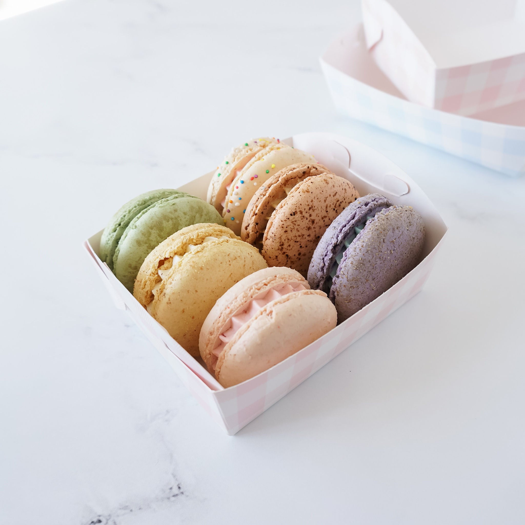 Six pastel colored macarons in a pink and white gingham box. The macarons are arranged in a row, with the green macaron on the left and the purple macaron on the right. The macarons are all different colors and have a variety of toppings.