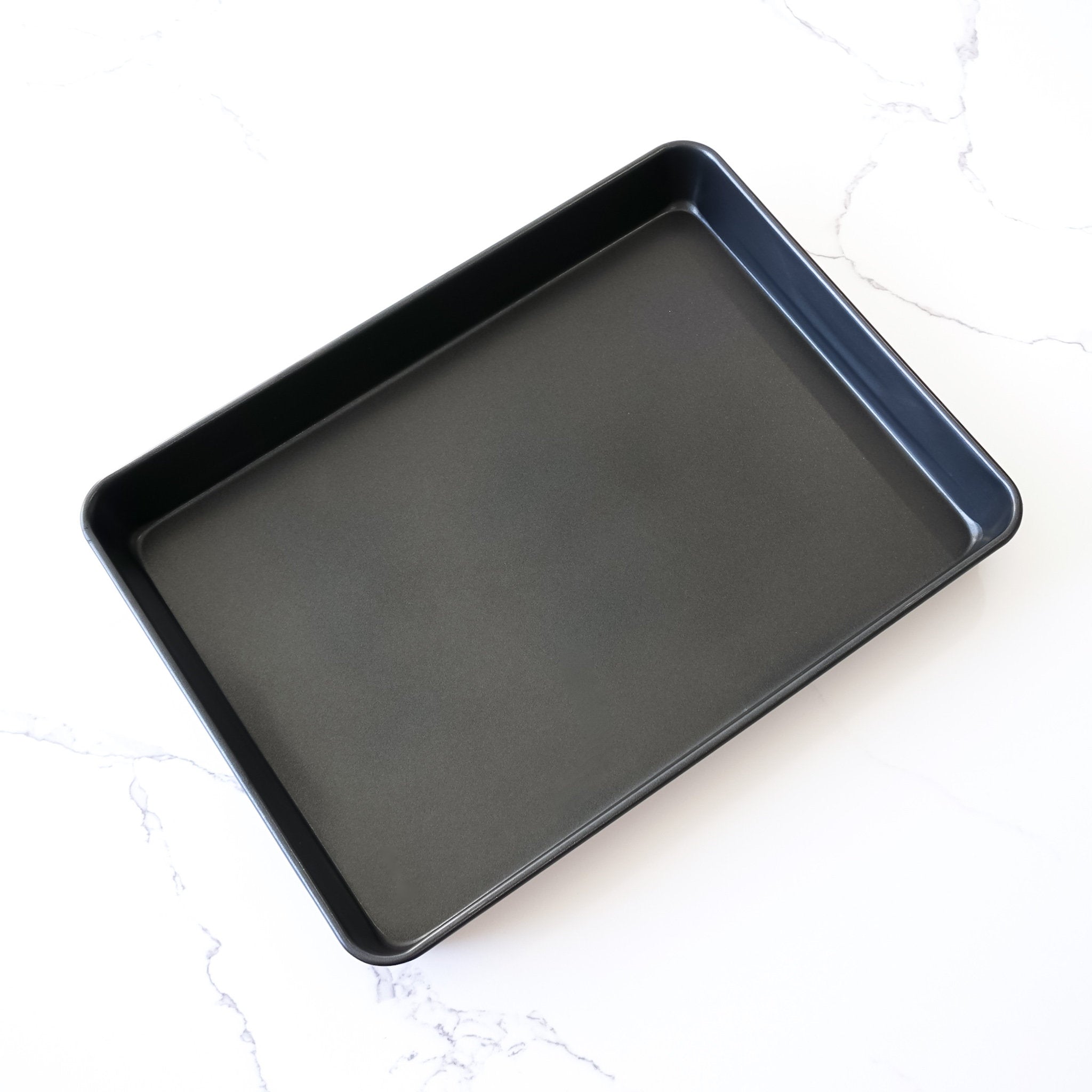 A rectangular, black sheet cake pan made of nonstick coating material, placed on a white marble surface.