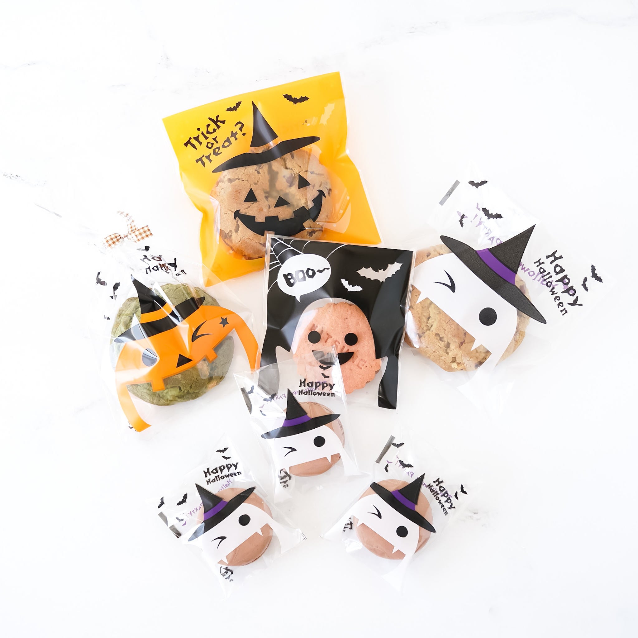 Assorted Halloween-themed cellophane bags with various designs including pumpkin, bat, and witch motifs, containing cookies or macarons.