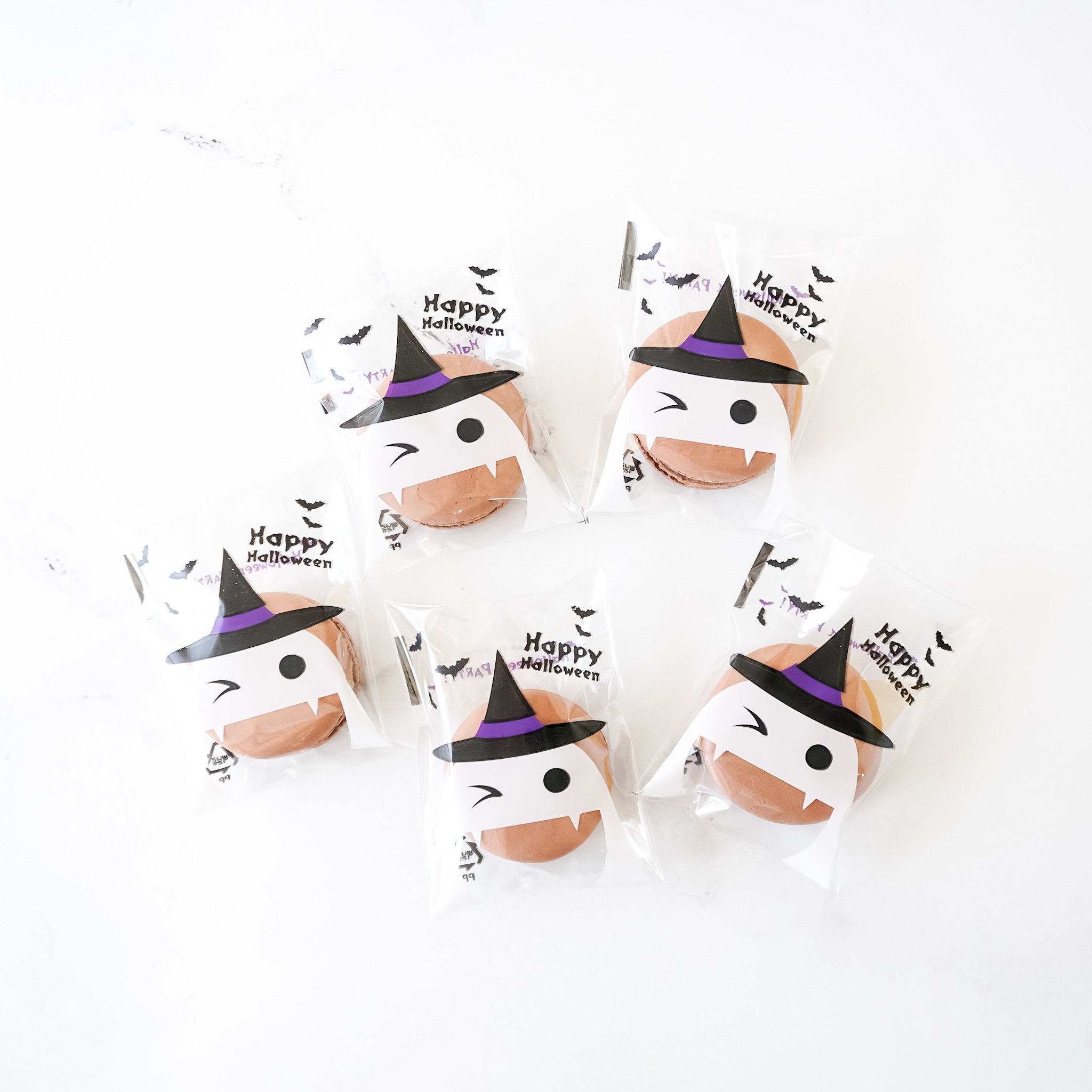 Halloween-themed cellophane bags with various designs including pumpkin, bat, and witch motifs, containing cookies or macarons.