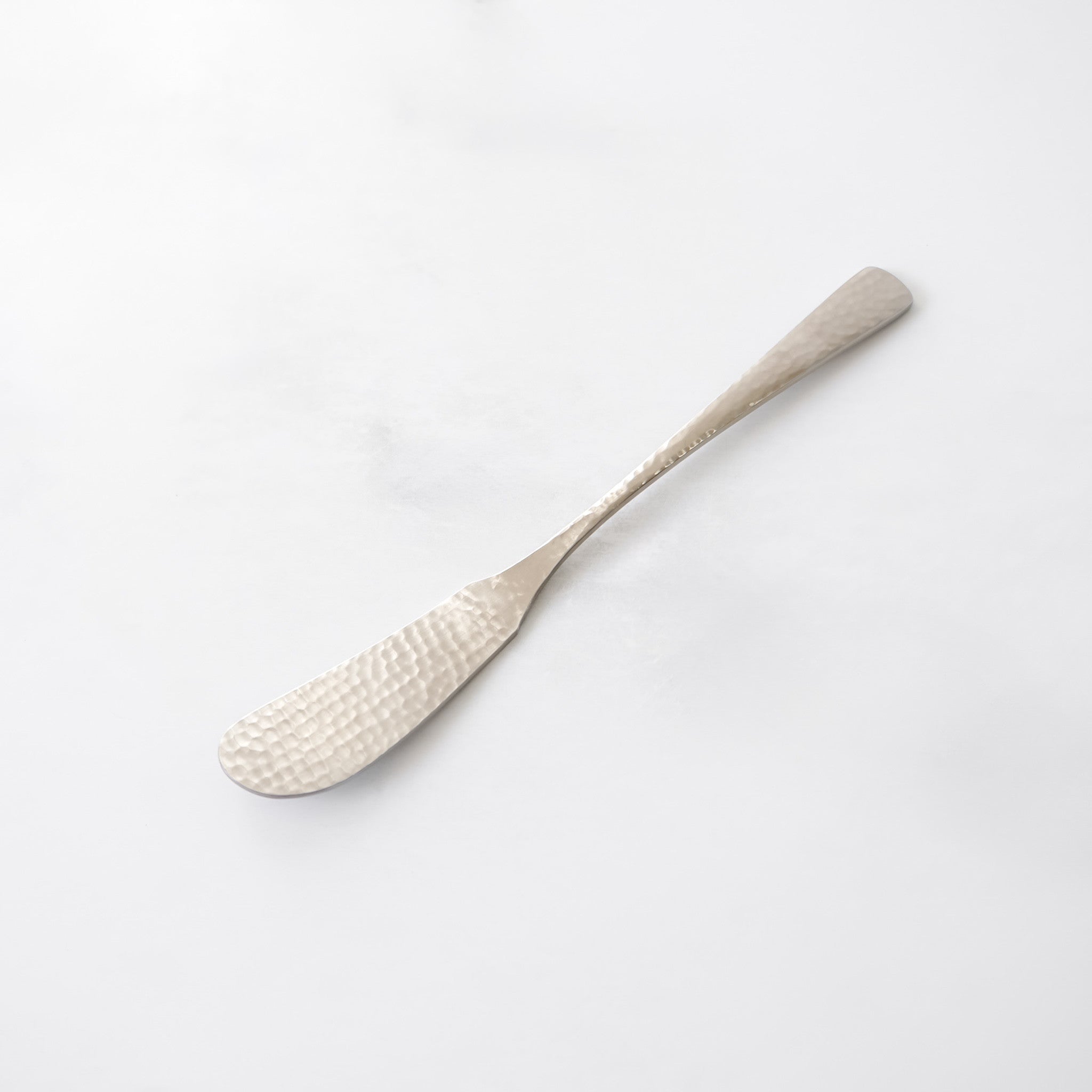 Silver butter knife with a textured, hammered design on the blade. The handle is slender and tapers to a rounded end. The knife is made of a shiny, metallic material and is set against a white background.