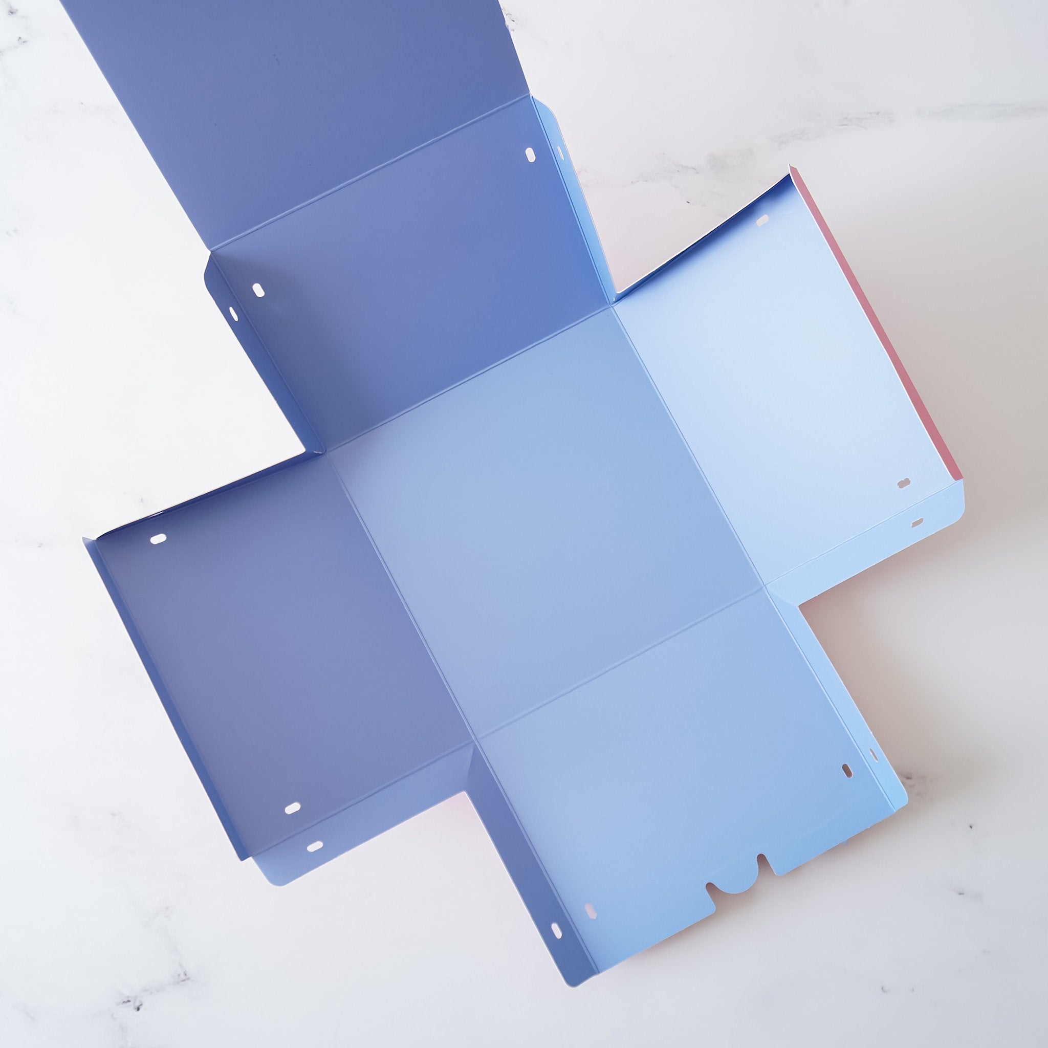 A blue and pink paper box with a square base and four flaps. The flaps are folded down and the box is open, revealing the inside of the box. The box has small holes in the flaps.