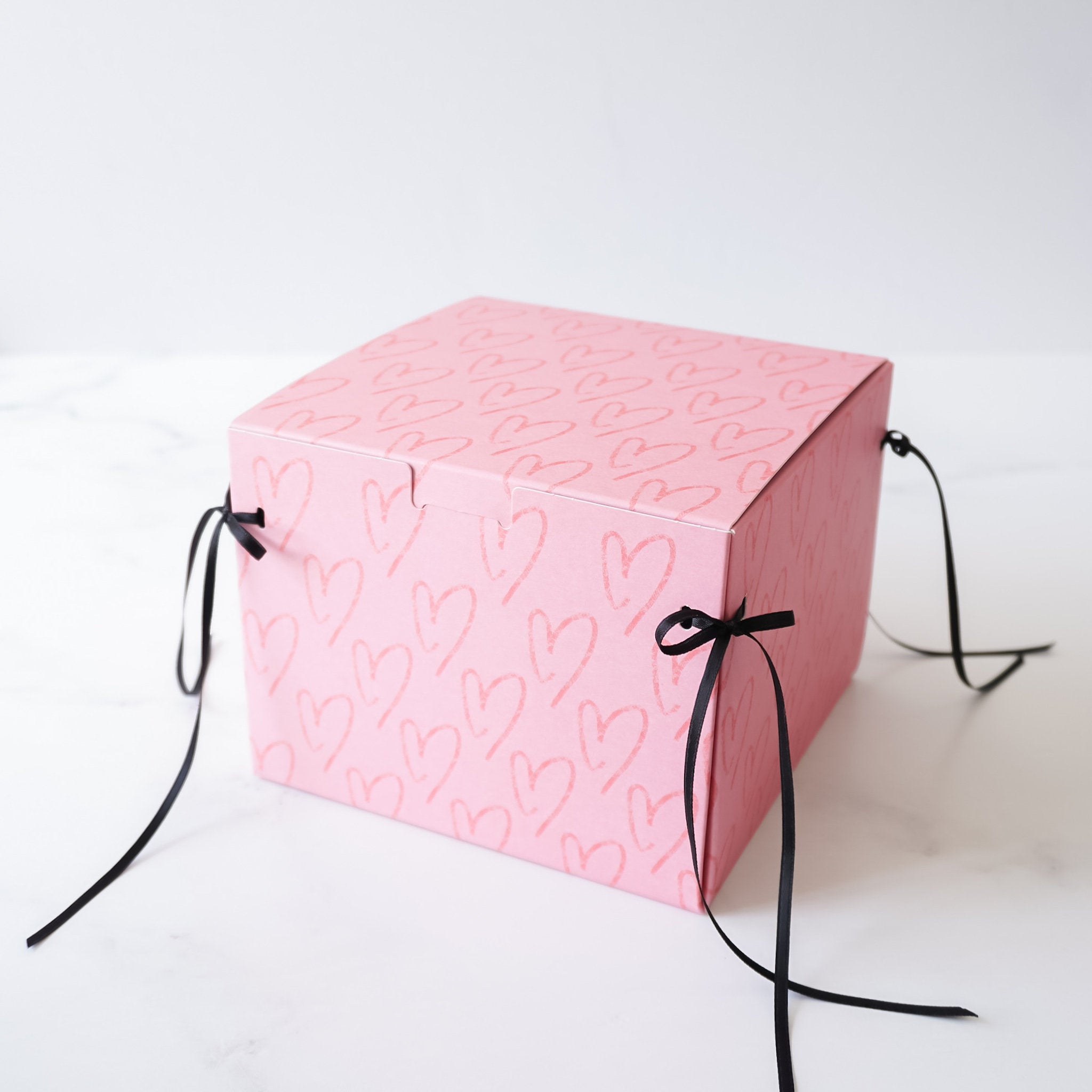A pink box with a heart pattern and black ribbon ties. The box is closed and the ribbon ties are tied in a bow.
