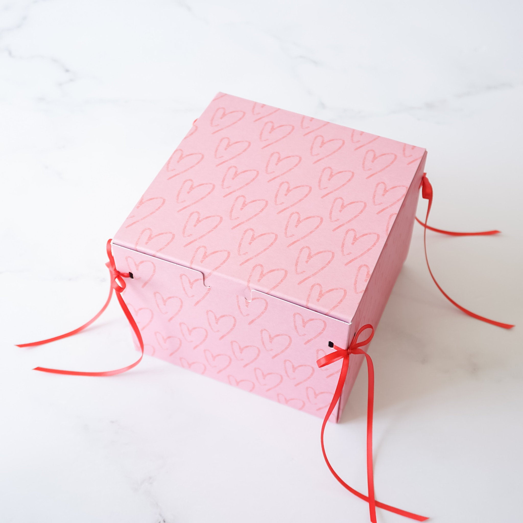 A pink gift box with a heart pattern and red ribbon ties. The box is empty and ready to be filled with a gift.
