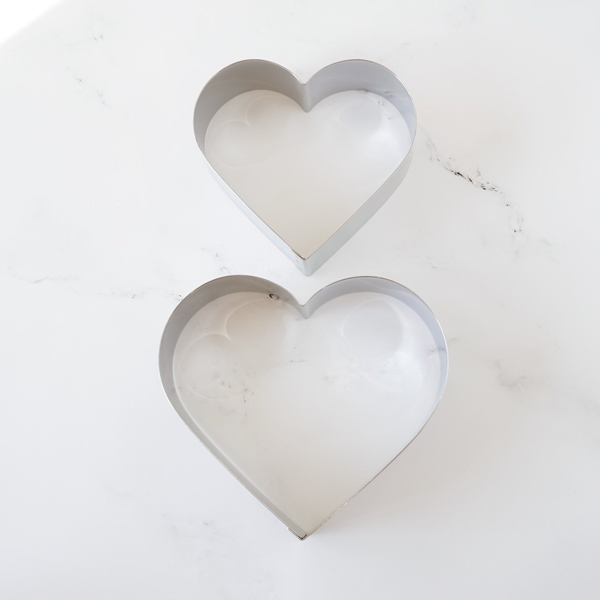 Two silver heart-shaped cake rings on a white marble surface. The larger cutter is on top of the smaller cutter.