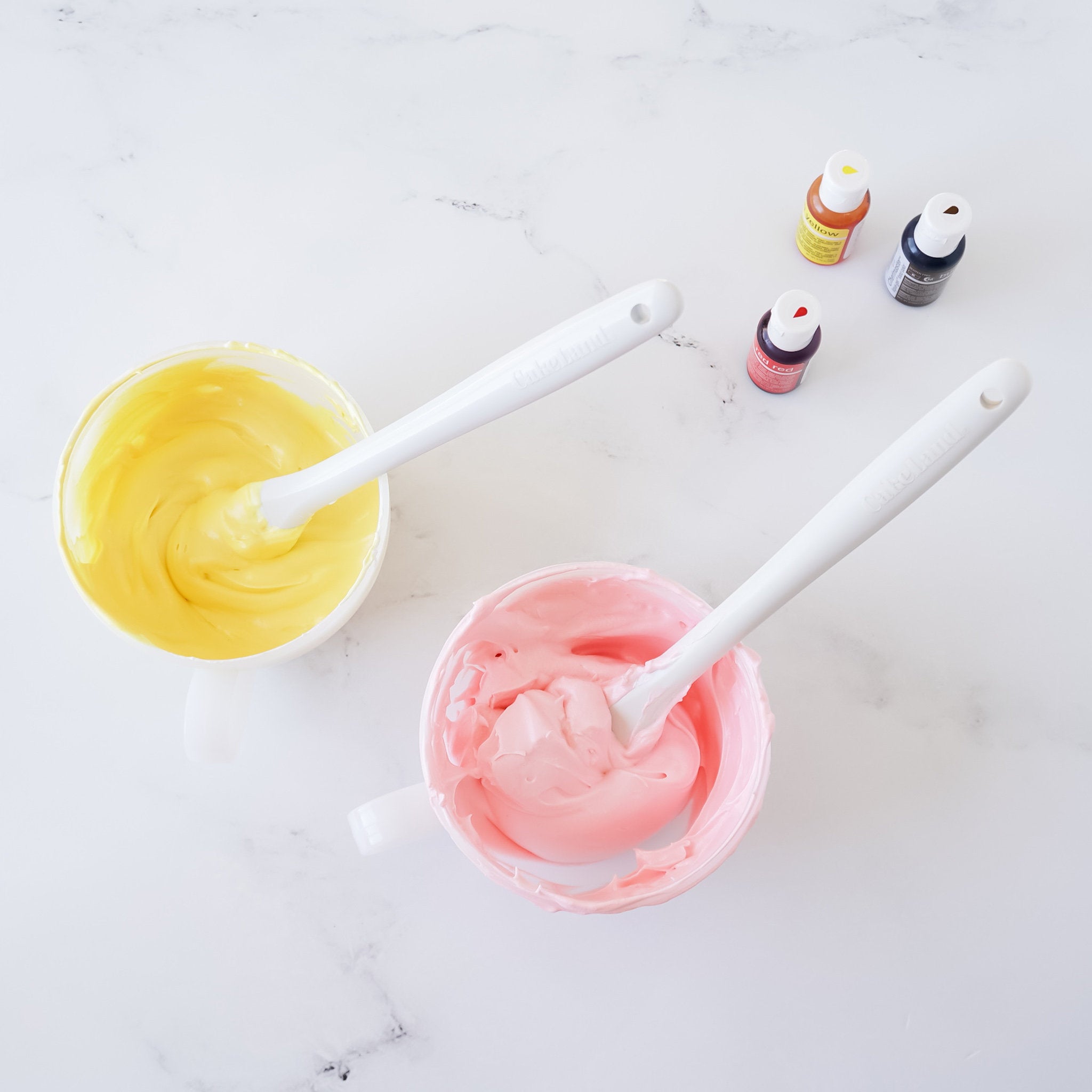 Two white bowls with yellow and pink frosting inside. Two white spatulas are in each bowl. Three small bottles of food coloring are on a white surface.