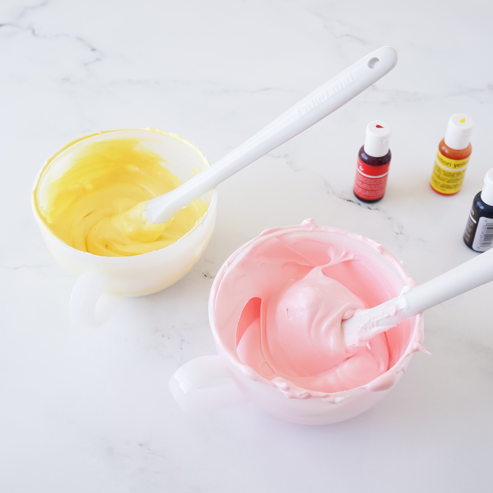 Two white bowls with yellow and pink frosting inside. A white spatula is in each bowl. Two bottles of red and yellow food coloring are in the background.