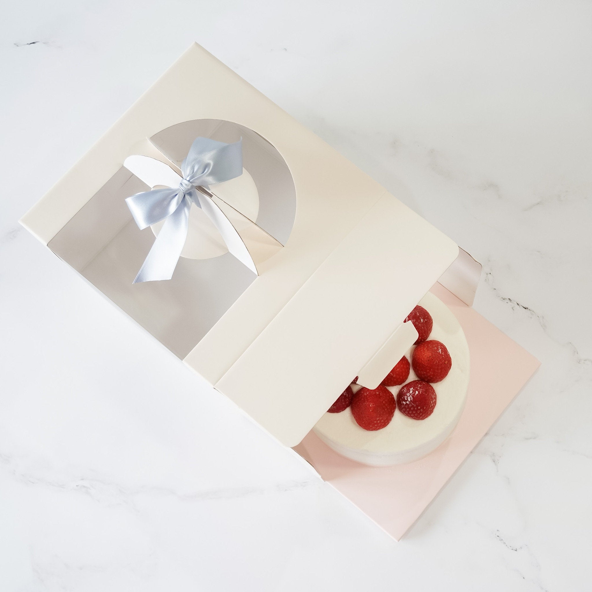 An ivory cake box with a window, open and unassembled, displaying a cake with strawberries inside, tied with a blue ribbon.
