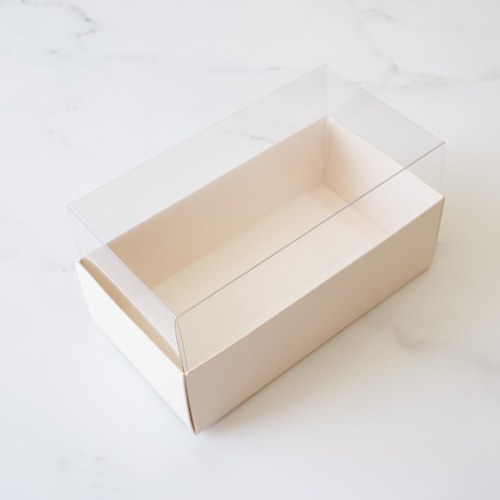 A rectangular cardboard box with a clear plastic lid. The box is an ivory color and is empty.