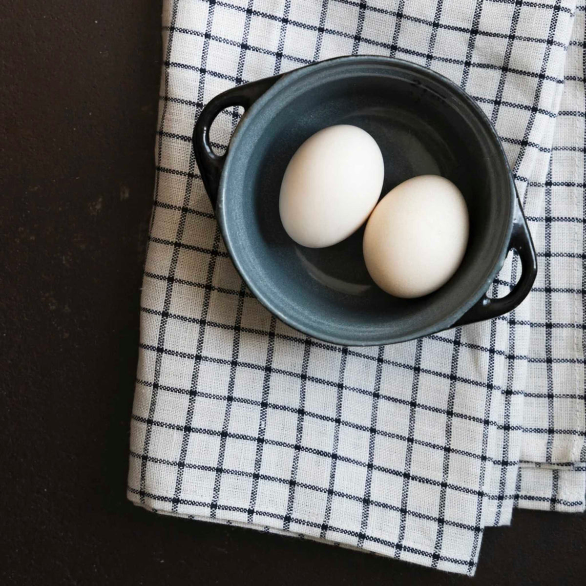 Two white eggs resting in a small dark gray ceramic pot with side handles, placed on a black-and-white checkered cloth over a dark surface.