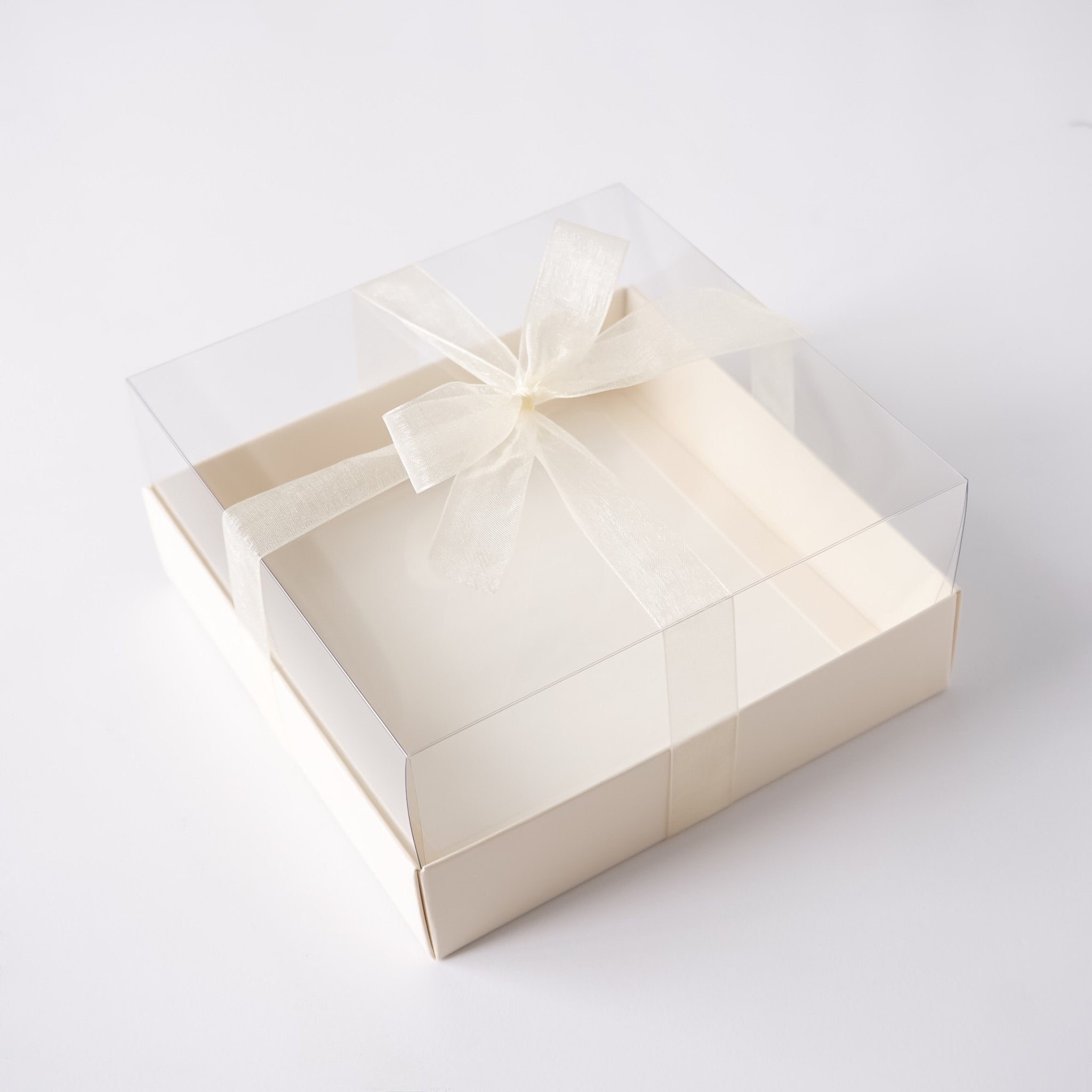 A square cream-colored bakery box with a clear plastic lid and a sheer cream ribbon tied in a bow, photographed on a clean white background.