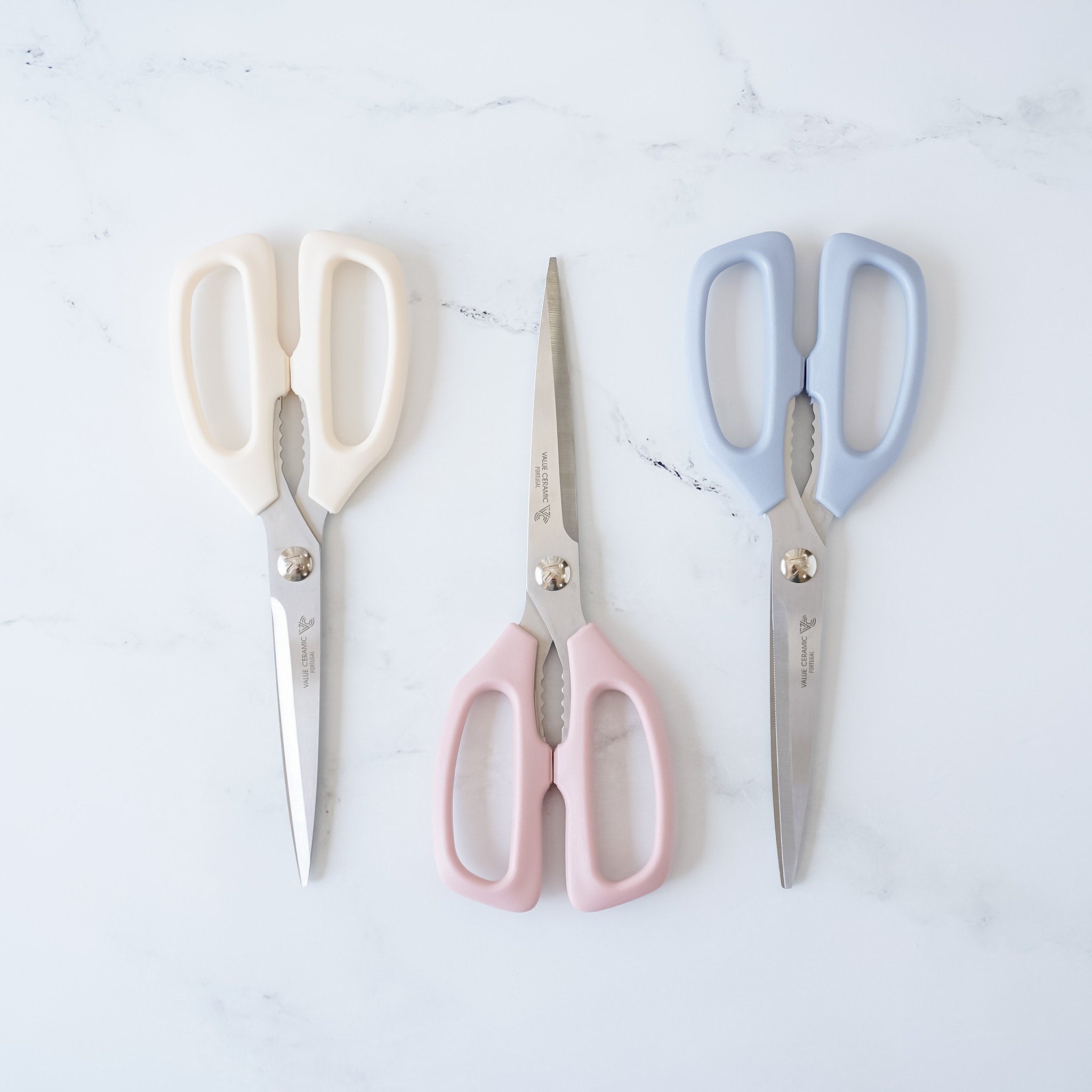 Three kitchen shears with stainless steel blades and ergonomic plastic handles, displayed in different colors (ivory, pink, and blue) on a marble surface.