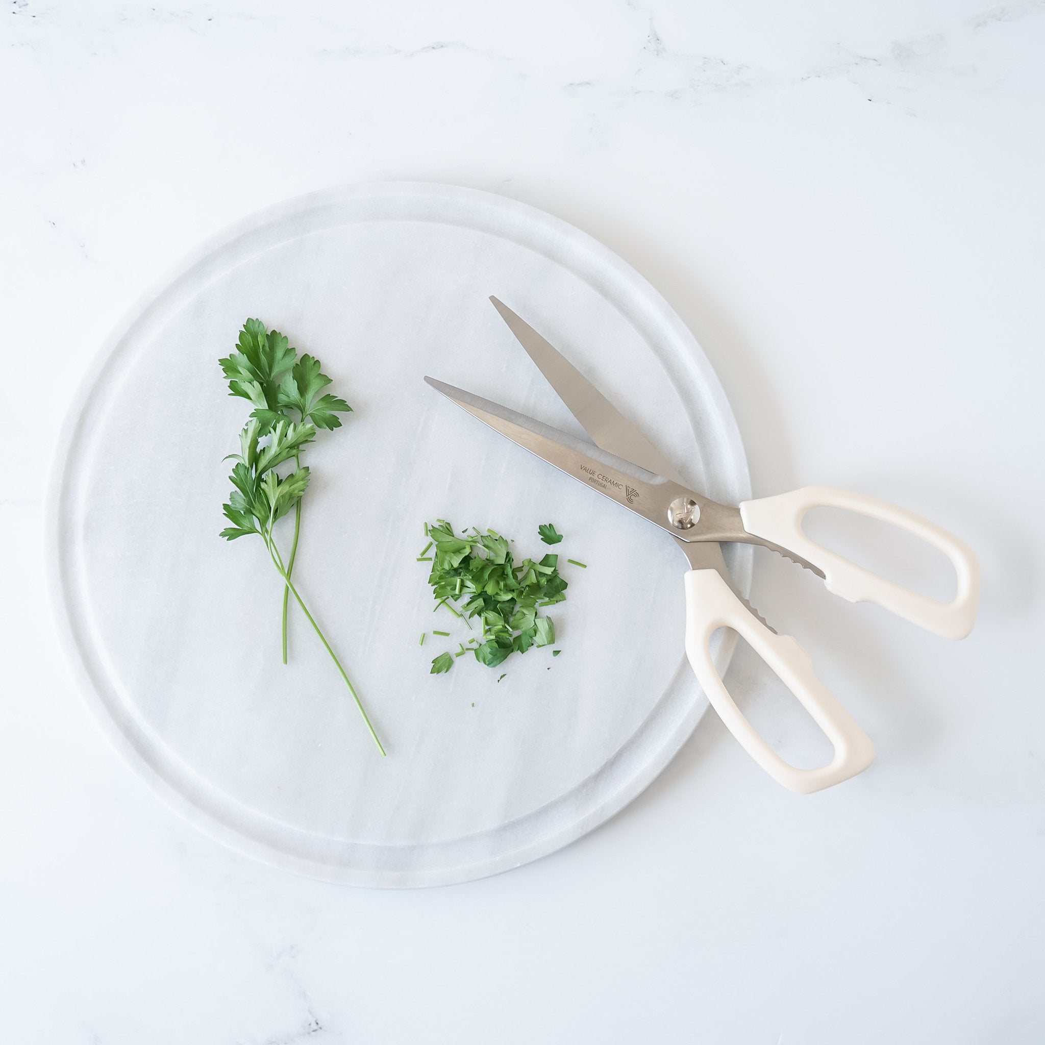 A white round cutting board with a sprig of parsley and chopped parsley. A pair of white handled kitchen shears are on the cutting board.