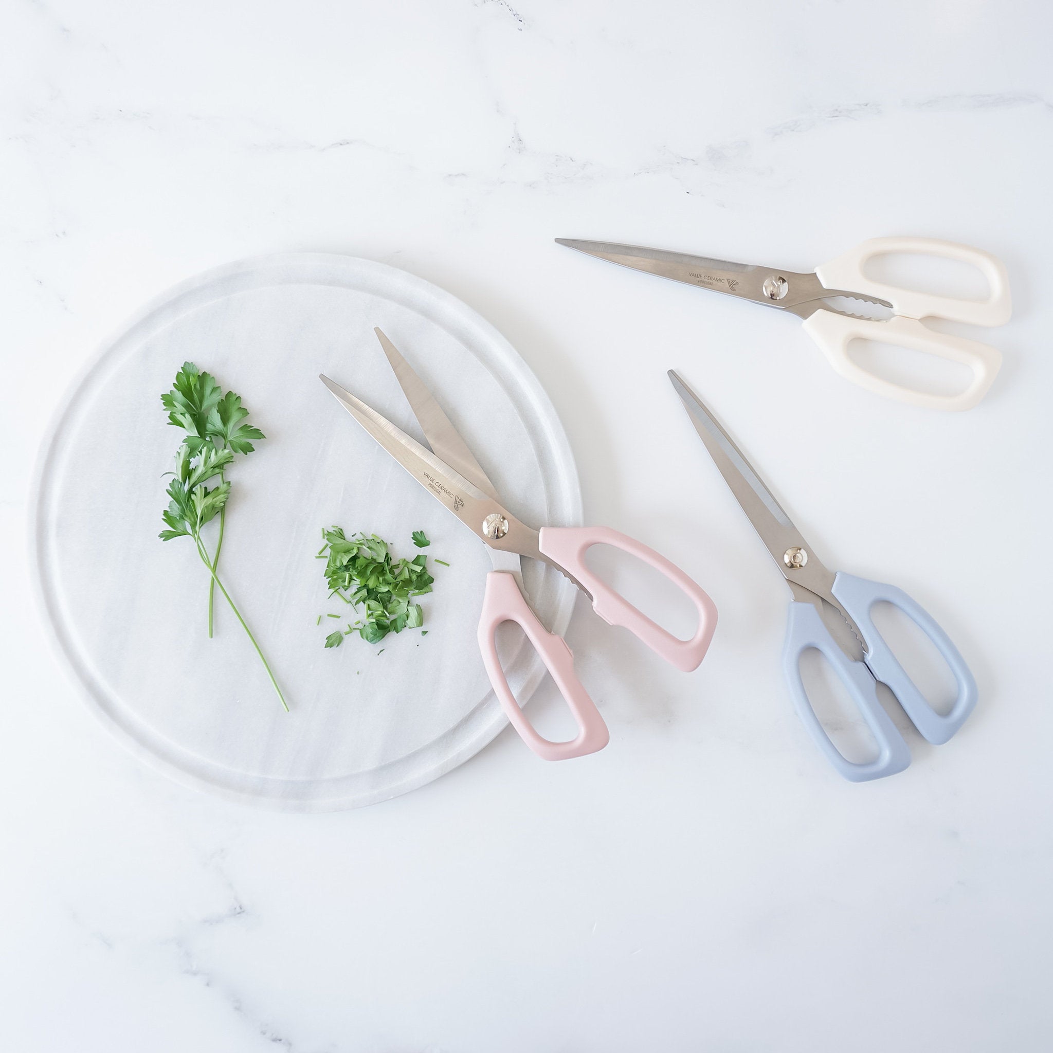 left have pink handles, the shears in the middle have white handles, and the shears on the right have blue handles. All three pairs of shears are on a white marble surface.