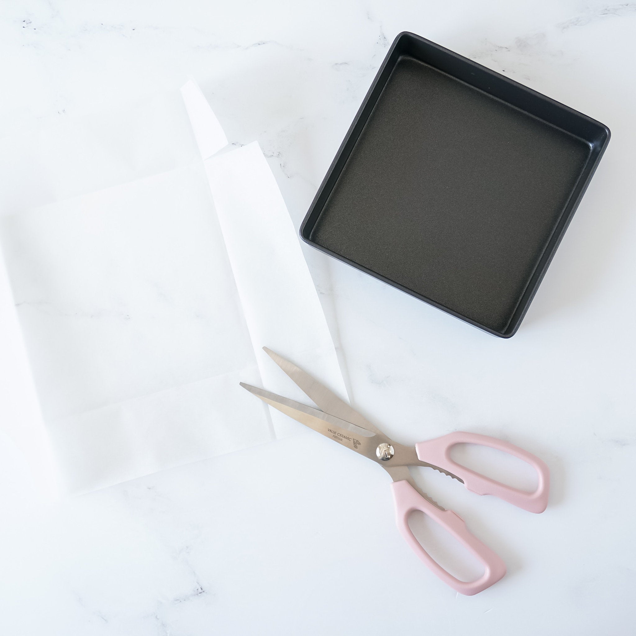 A pair of pink handled kitchen shears with a stainless steel blade. The shears are lying on a white surface next to a white parchment paper and a black square baking pan.
