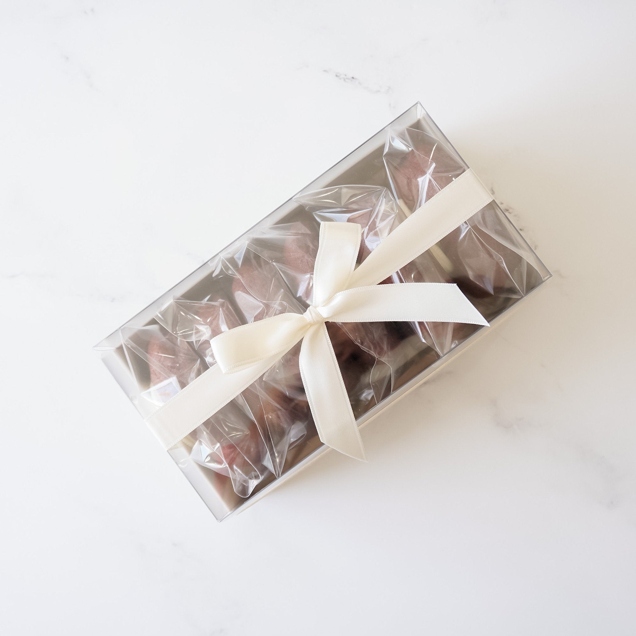 A clear plastic box with a white ribbon tied around it. The box contains six small, brown chocolate treats.