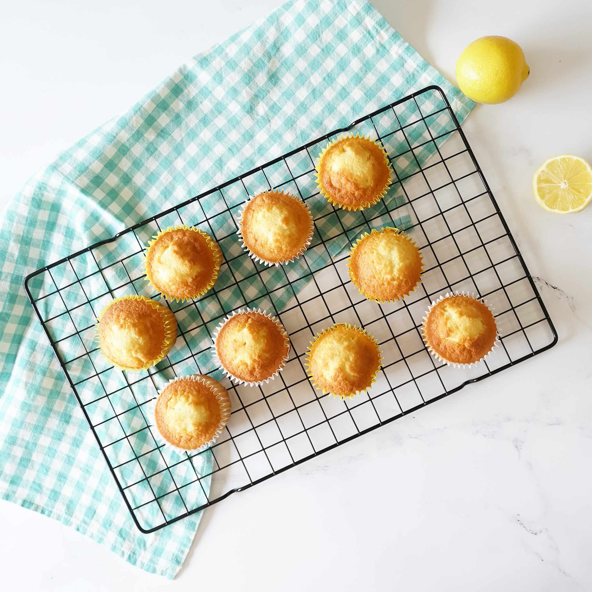 A black wire rack with nine yellow lemon cupcakes on it. The cupcakes are arranged in a circle on the rack. The rack is on top of a green and white checkered tablecloth.