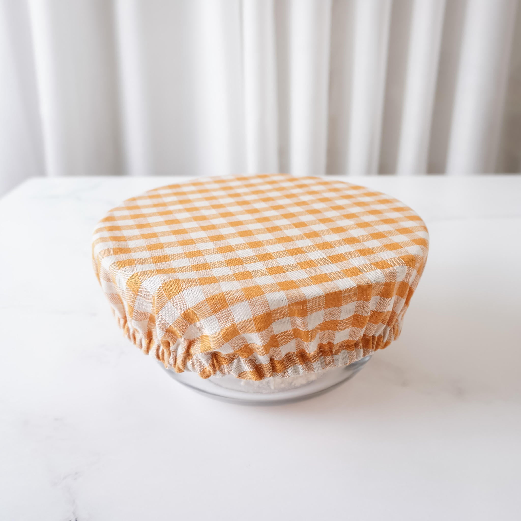 A bowl cover in orange and white checkered pattern, sized large, displayed on a white surface with a draped white curtain in the background.