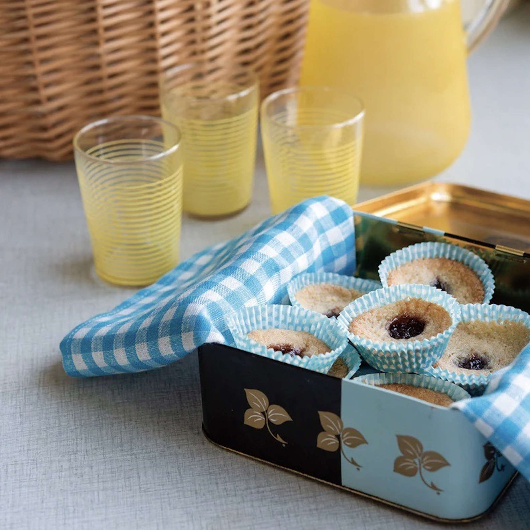A blue and white checkered linen dishcloth is laid inside a tin case, topped with baked goods. In the background, three yellow-striped glasses sit empty beside a jar of orange juice.