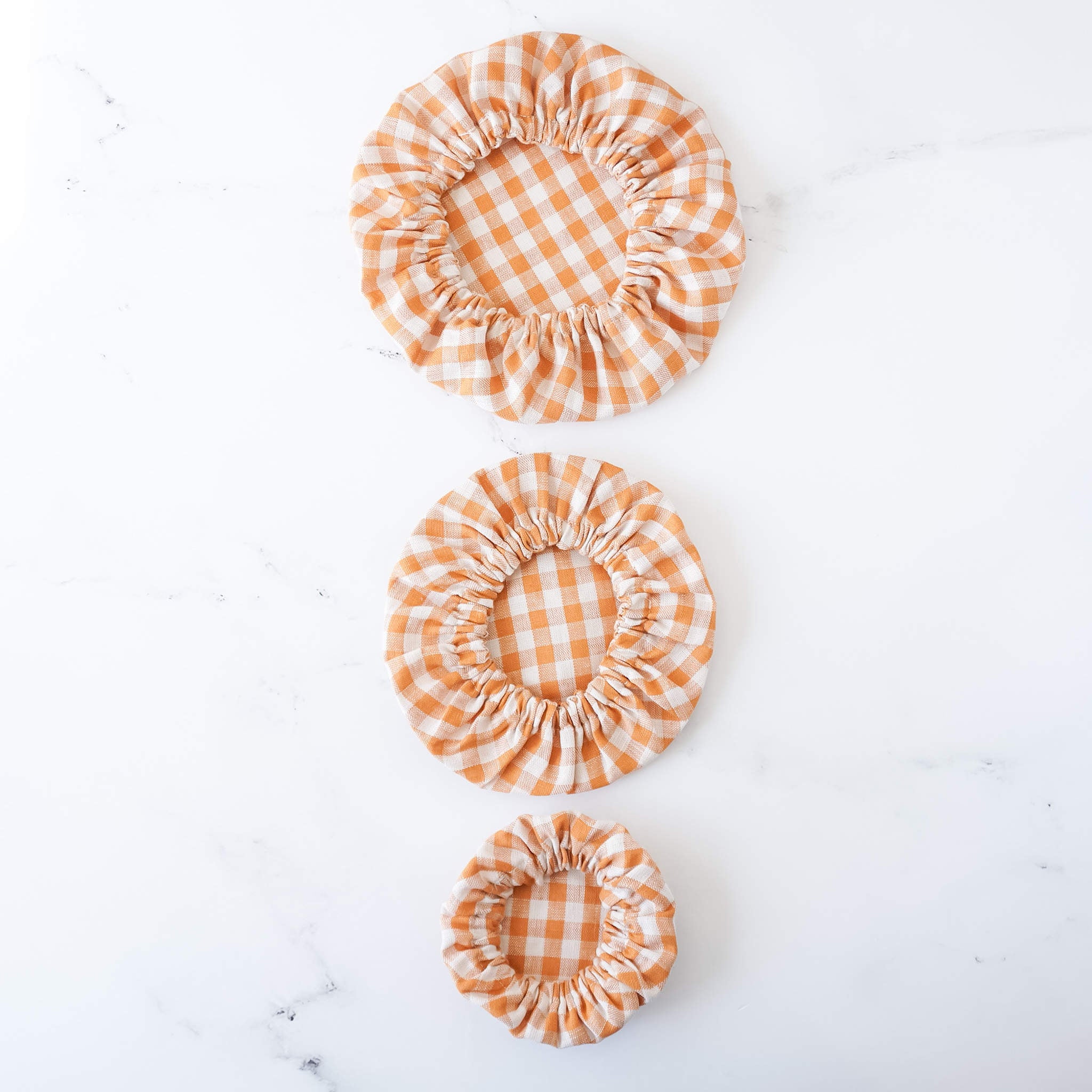 Three bowl covers in orange and white checkered pattern, sized small, medium, and large, displayed on a white surface.