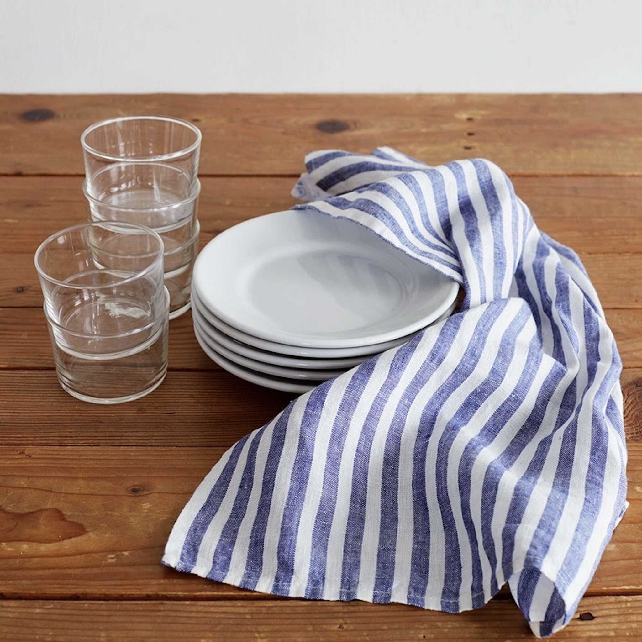 A blue and white striped linen dishcloth on a wooden table, with two clear glasses and a stack of white plates in the background.
