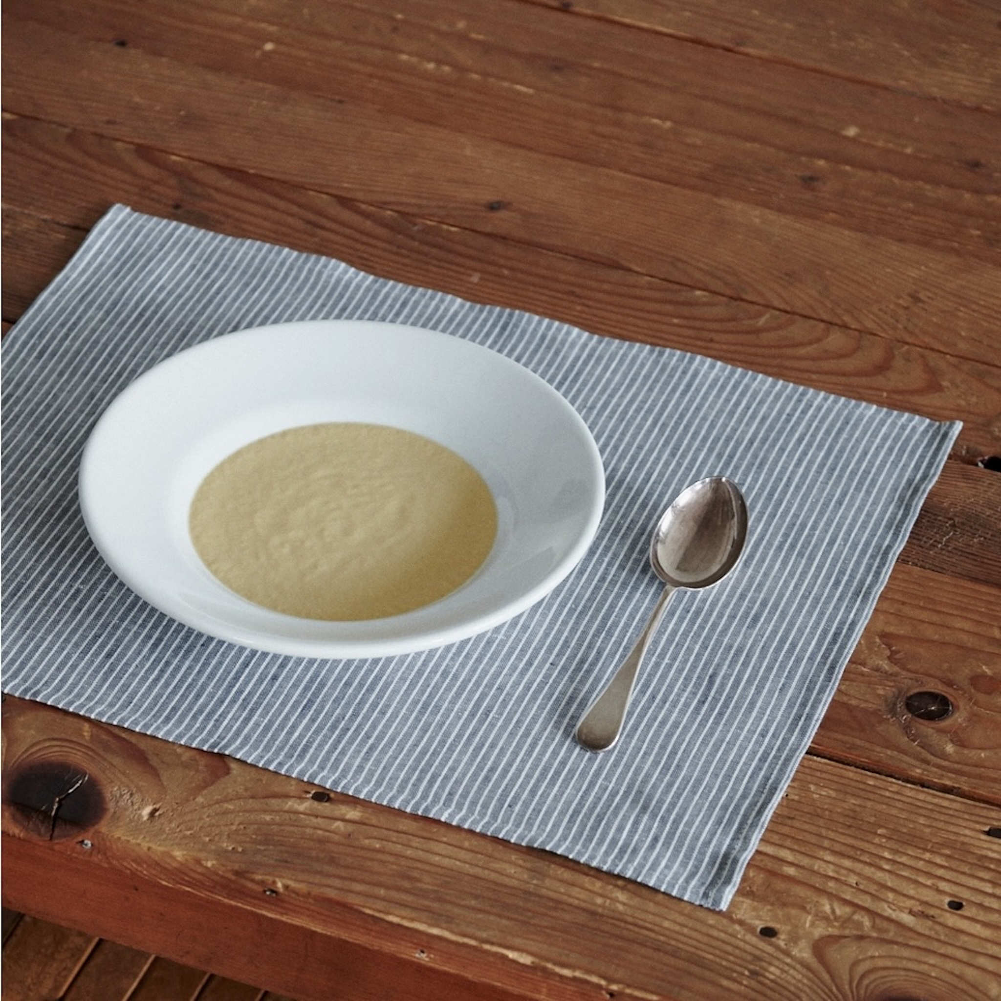 White plate with soup and a spoon on a gray striped placemat on a wooden table