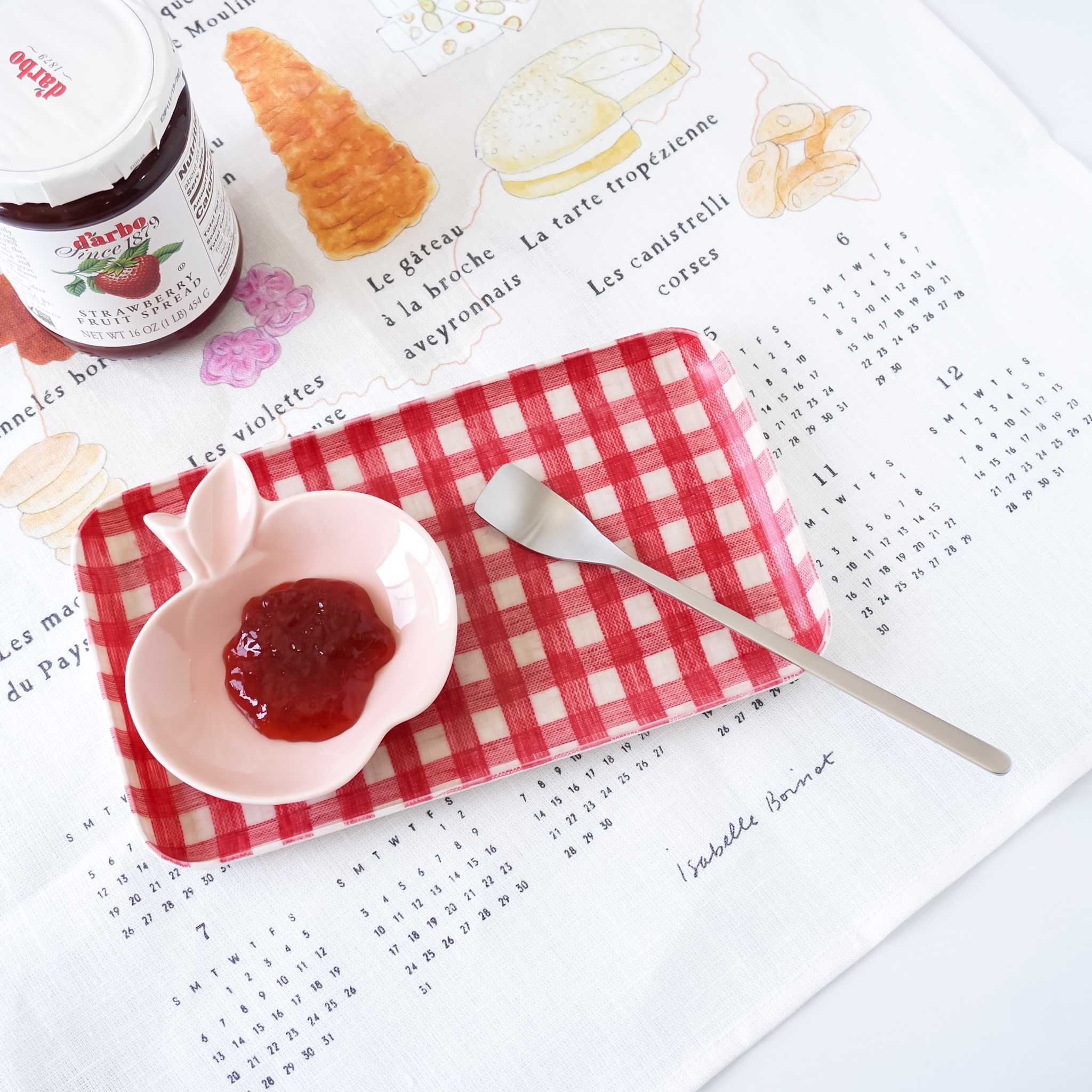 Small pink dish with jam, a spoon, and a jar of jam on a red checkered tray over printed kitchen cloth.