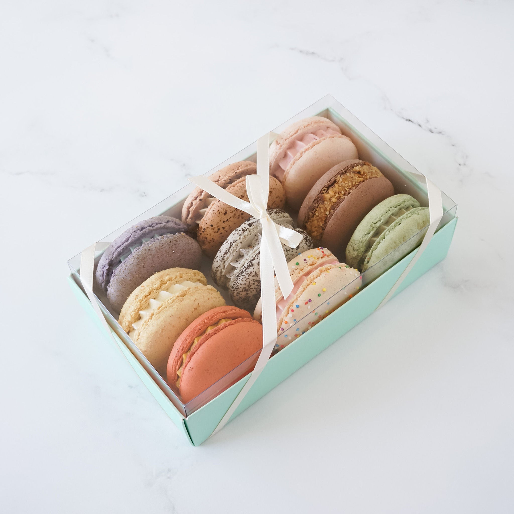 A box of assorted macarons in a mint green box with a white ribbon. The macarons are in various colors including pink, purple, green, brown, beige, and orange. The macarons are arranged in three rows of three.