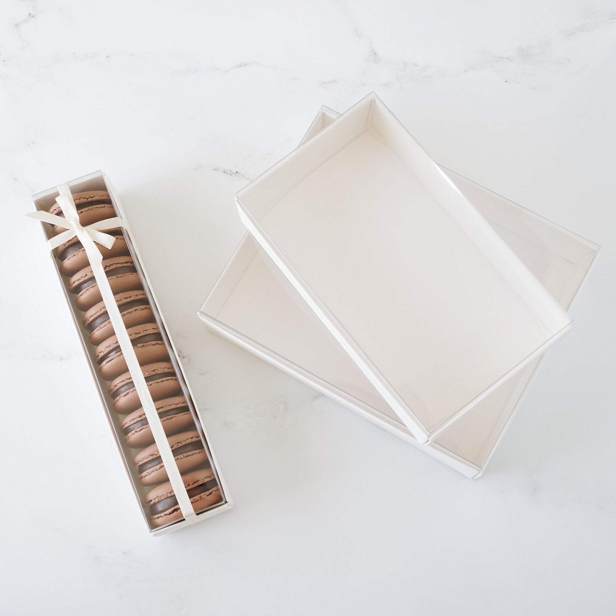 A white box with a clear lid containing a row of chocolate macarons tied with a white ribbon. The macarons are arranged in a row and are stacked on top of each other. The macarons are a light brown color and have a slightly glossy finish.