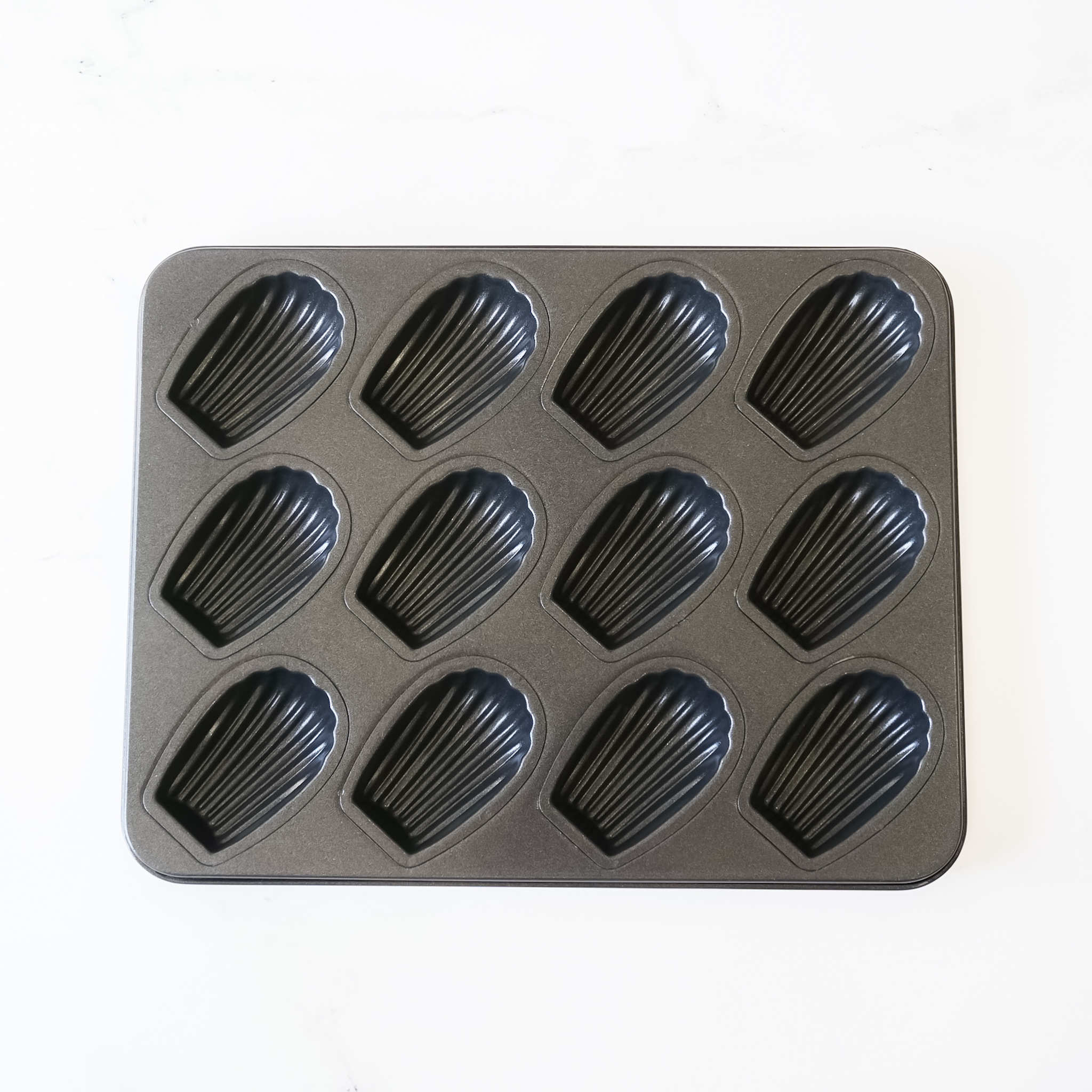 A black metal baking pan with 12 individual molds shaped like seashells. The pan is designed for making madeleines.