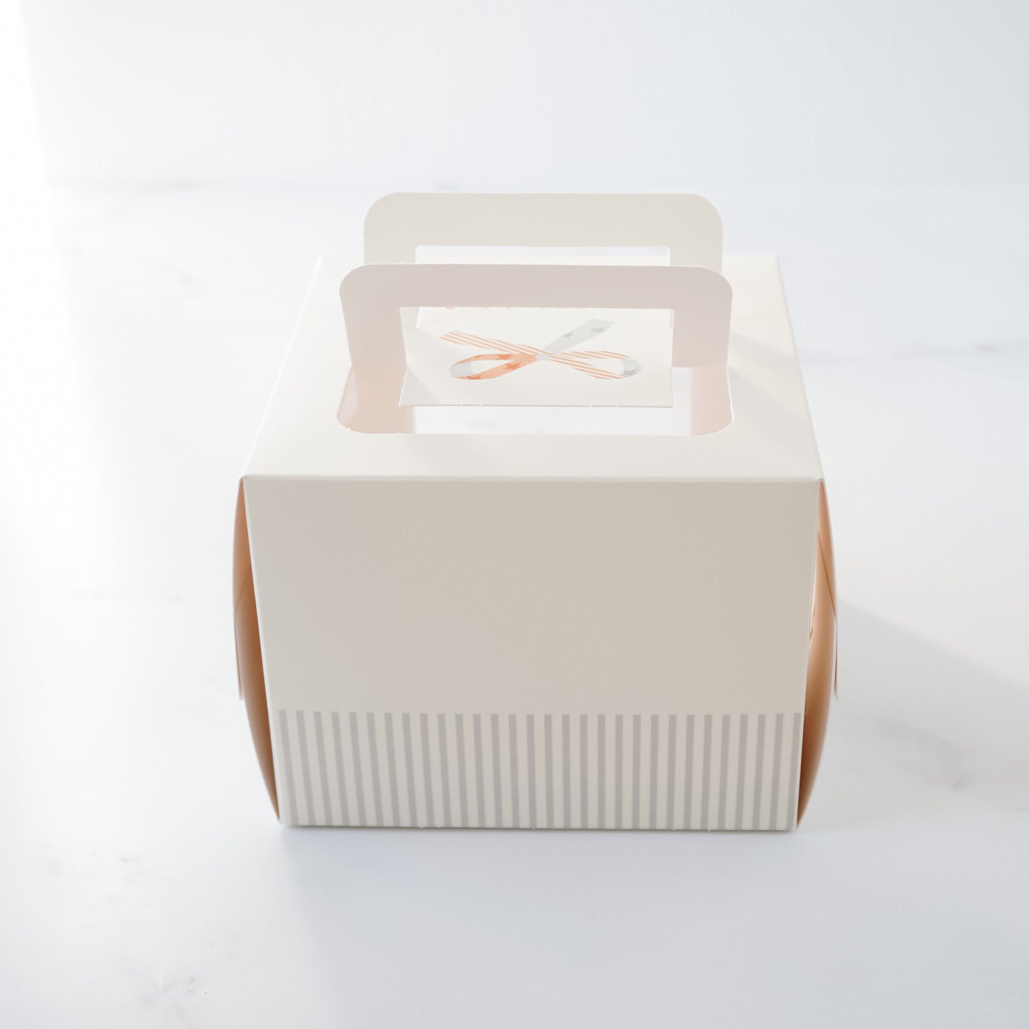 A white cardboard cake box with a window on the top. The window has a decorative brown and white ribbon design. The box has a striped pattern on the bottom and two handles on the sides.
