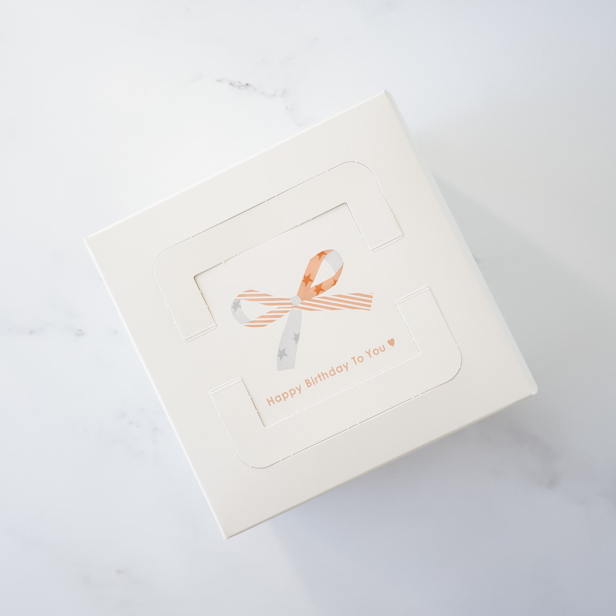 A white box with a cut-out window showing a pink and white ribbon bow with stars. The text &