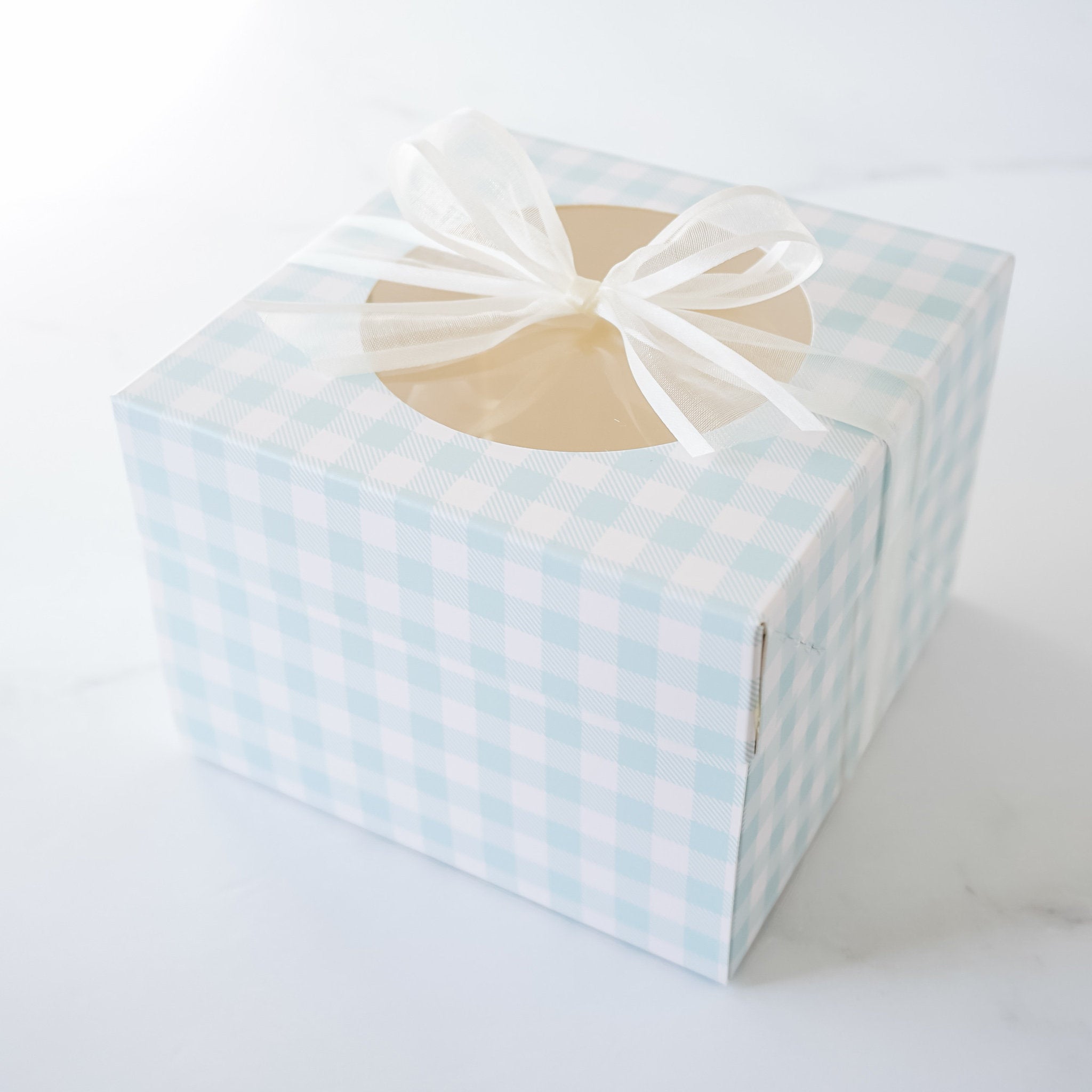 A light blue and white gingham gift box with a clear window and a white ribbon tied in a bow.