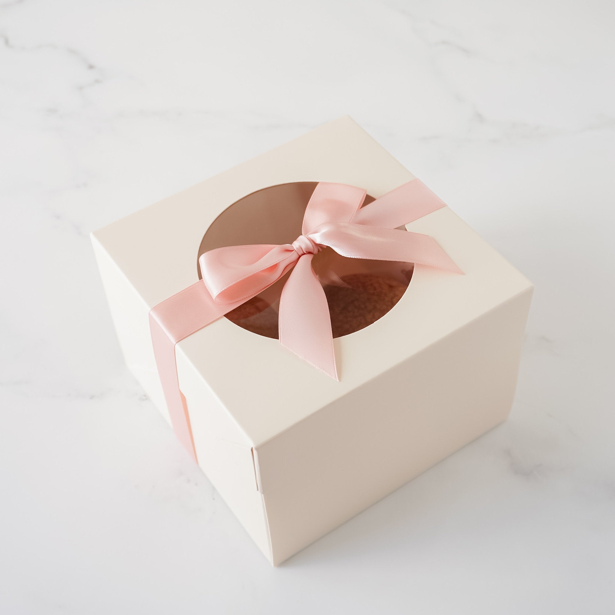 An ivory square box with a pink ribbon tied in a bow over a circular window. The window reveals a cake inside the box.
